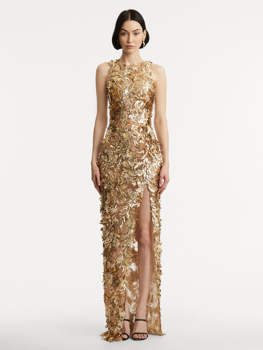 Full body front view of model wearing sleeveless gold-embroidered gown with a halter neckline and feathers unfurling across a tulle base. Gown features a high center front slit.