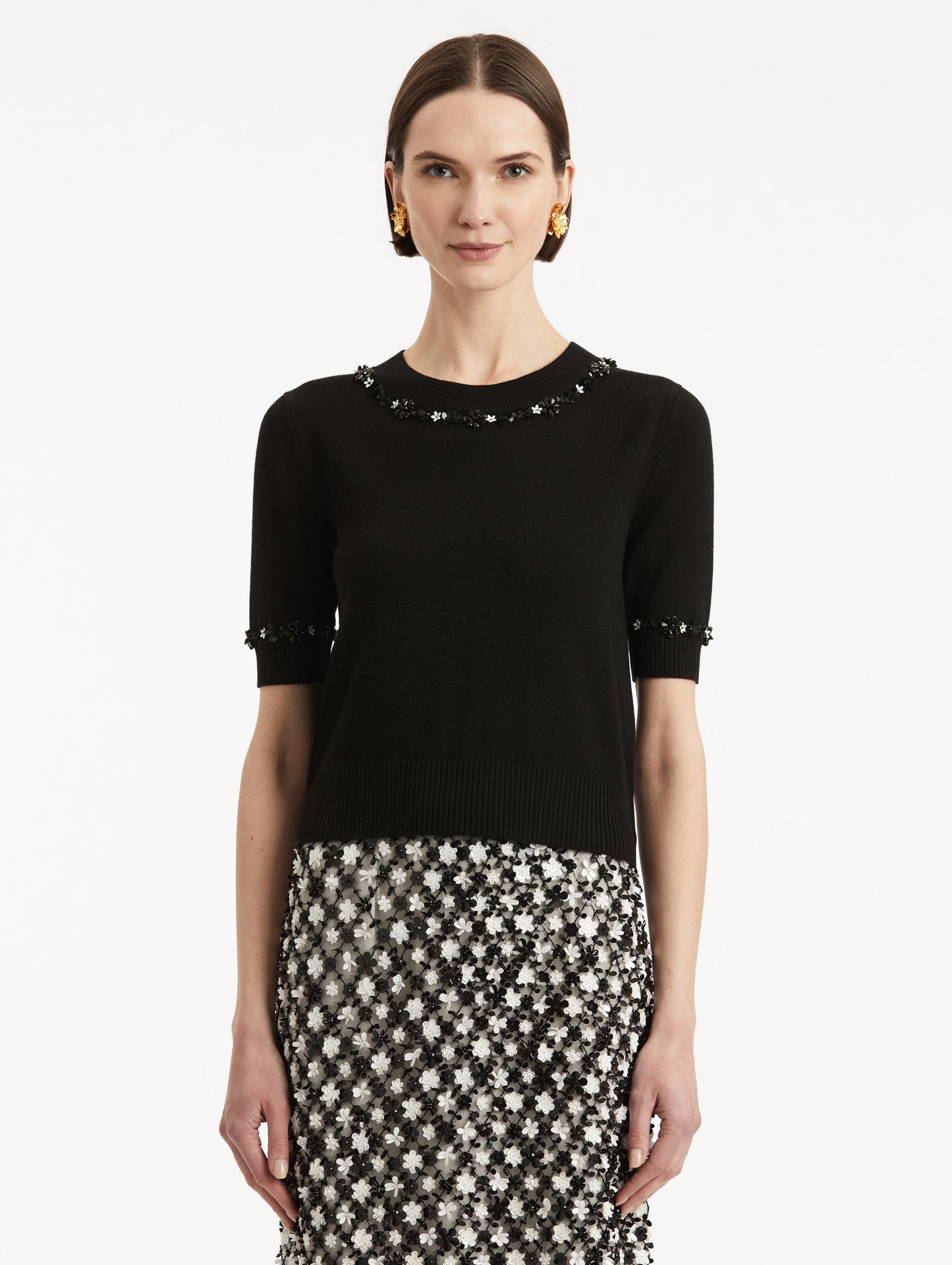 Front view of model wearing a short sleeve black pullover with a crew neckline and intricate sequin embroidery of ivory and noir florets which trace the trim.