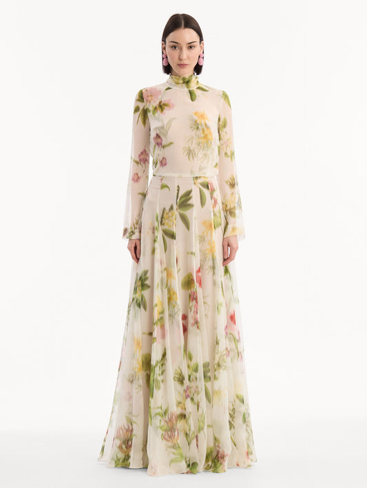 The model is wearing the Chiné Dominican Mixed-Floral Chiffon Gown in Ivory printed with an array of Dominican florals. Featuring long sleeves, mock neck, and flowing, pleated skirt. Front View.