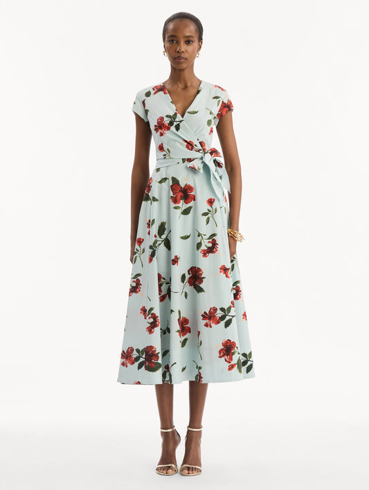 The model is wearing the Hibiscus Cotton Poplin Wrap Dress in light blue printed with red hibiscus blooms. Featuring a wrap construction bodice, cap sleeves, V-neckline, and full midi skirt. Front View.