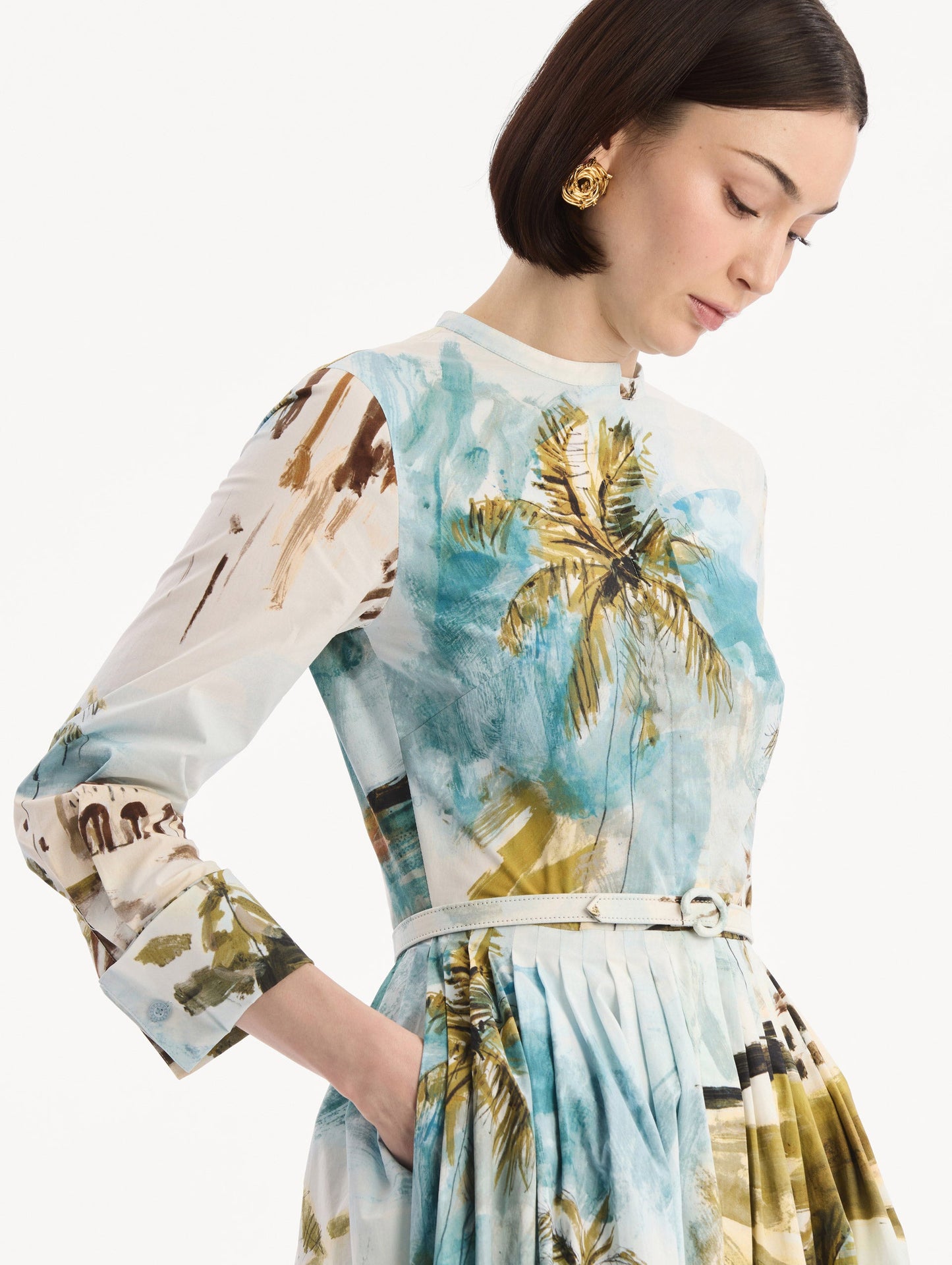 The model is wearing the Dominican Landscape Cotton Poplin Short-Sleeve Dress printed with the Dominican landscape. Featuring long sleeves, front-button closure, belt at waist, and pleated midi skirt. Detail View.