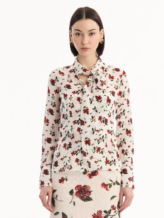 The model is wearing the Hibiscus Silk Chiffon Blouse in white printed with red hibiscus florals. Featuring a tie at neckline, long sleeves, and tailored silhouette. Front View.