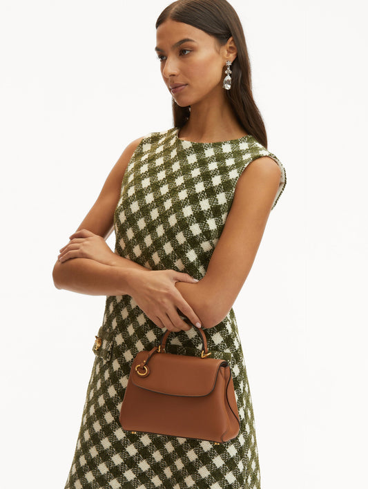 Model in plaid dress holding The Mini Poppy leather bag in brown with O logo around the top handle. Front View