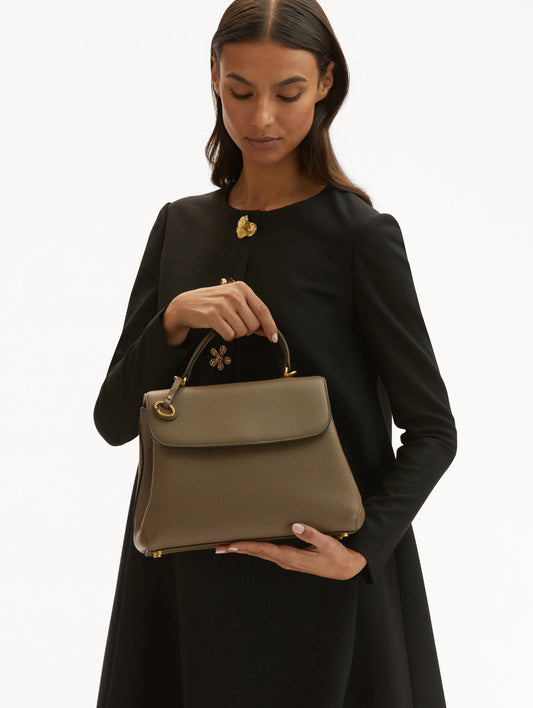 Model in black coat holding The Poppy leather handbag in taupe with O logo tag around the top handle. Front View