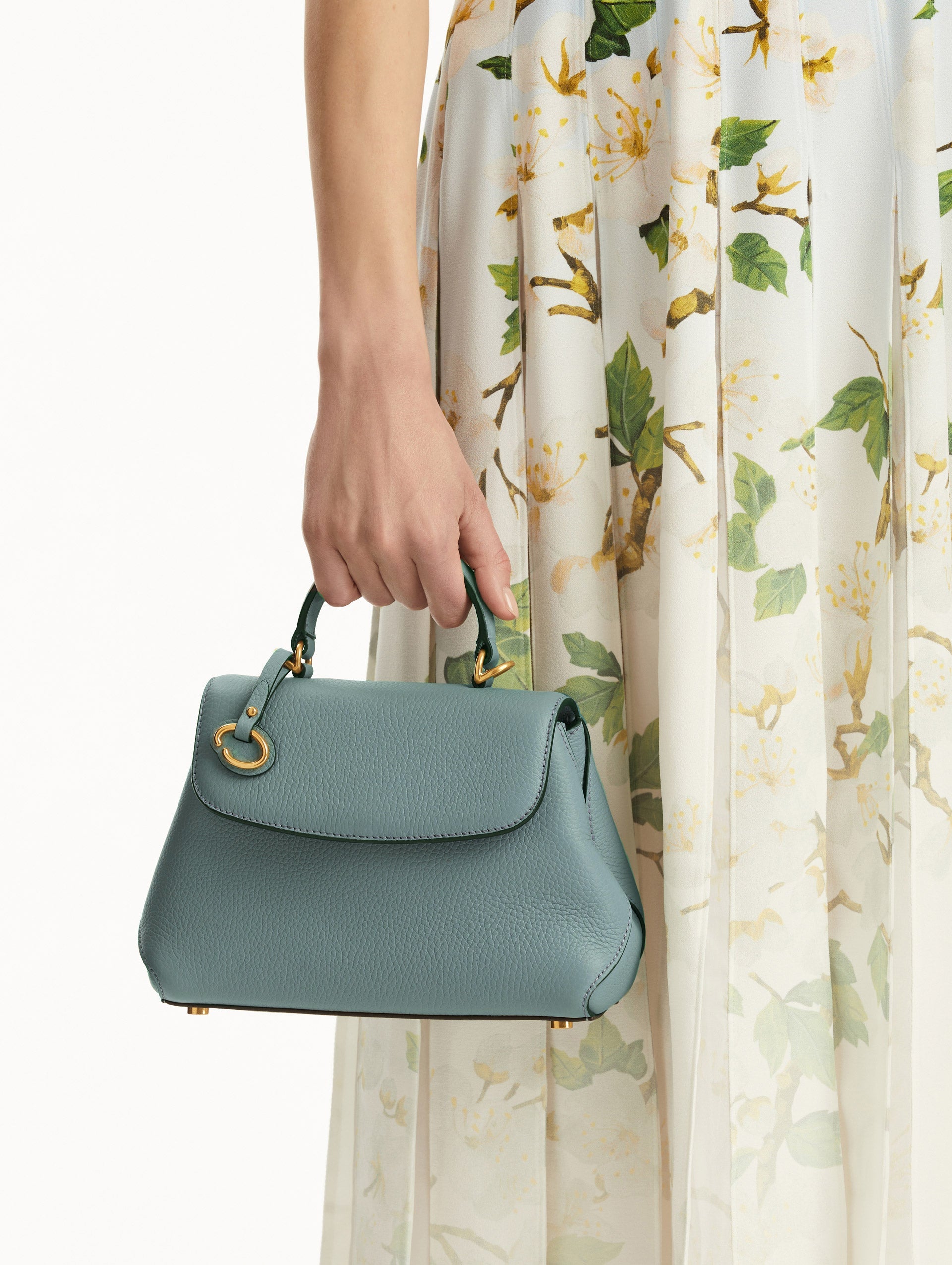 The model is holding the Mini Poppy handbag in a Mint color with an Oscar O logo tag around the top handle, paired with a floral white dress.
