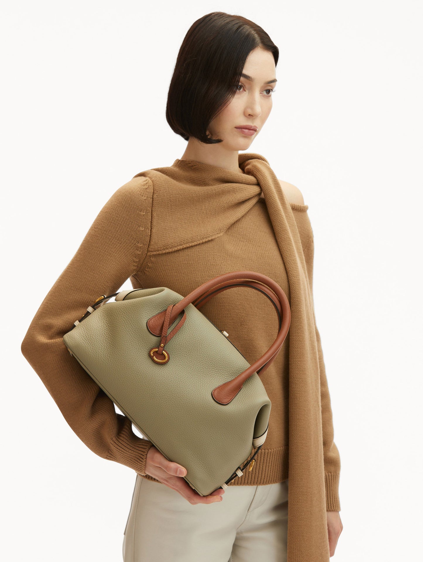 Model in Brown Sweater and White pants Wearing The Dahlia Duffle Bag in Sage Green with Brown Top Handle. Front View