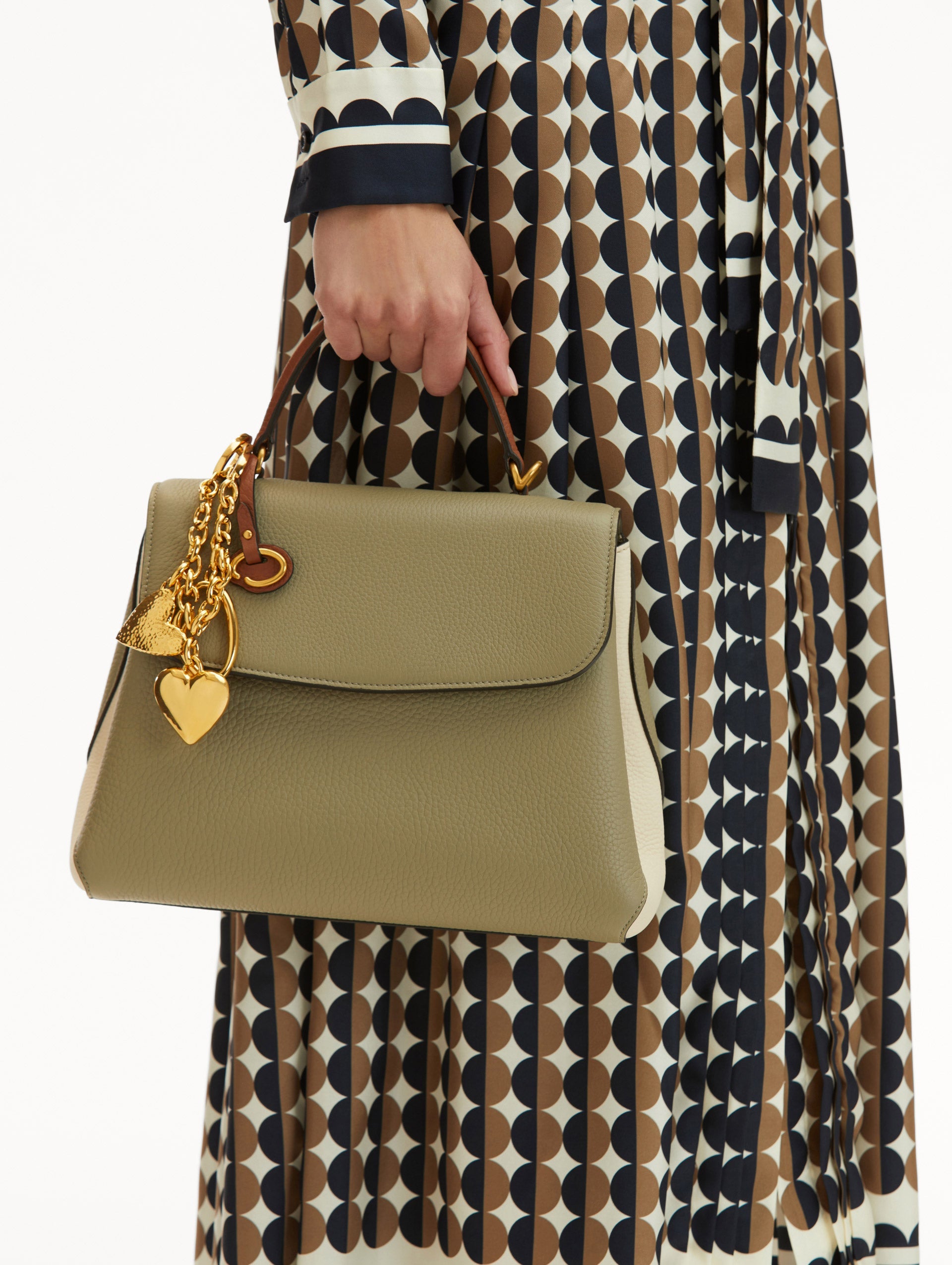 Model in Geometric Print dress wearing The Poppy Handbag in sage green with brown Top Handle. Adorned with Oscar de la Renta's signature O at the ornamental bag tag and hearts gold bag charm. Front View
