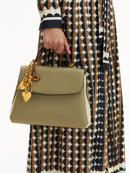 Model in Geometric Print dress wearing The Poppy Handbag in sage green with brown Top Handle. Adorned with Oscar de la Renta's signature O at the ornamental bag tag and hearts gold bag charm. Front View