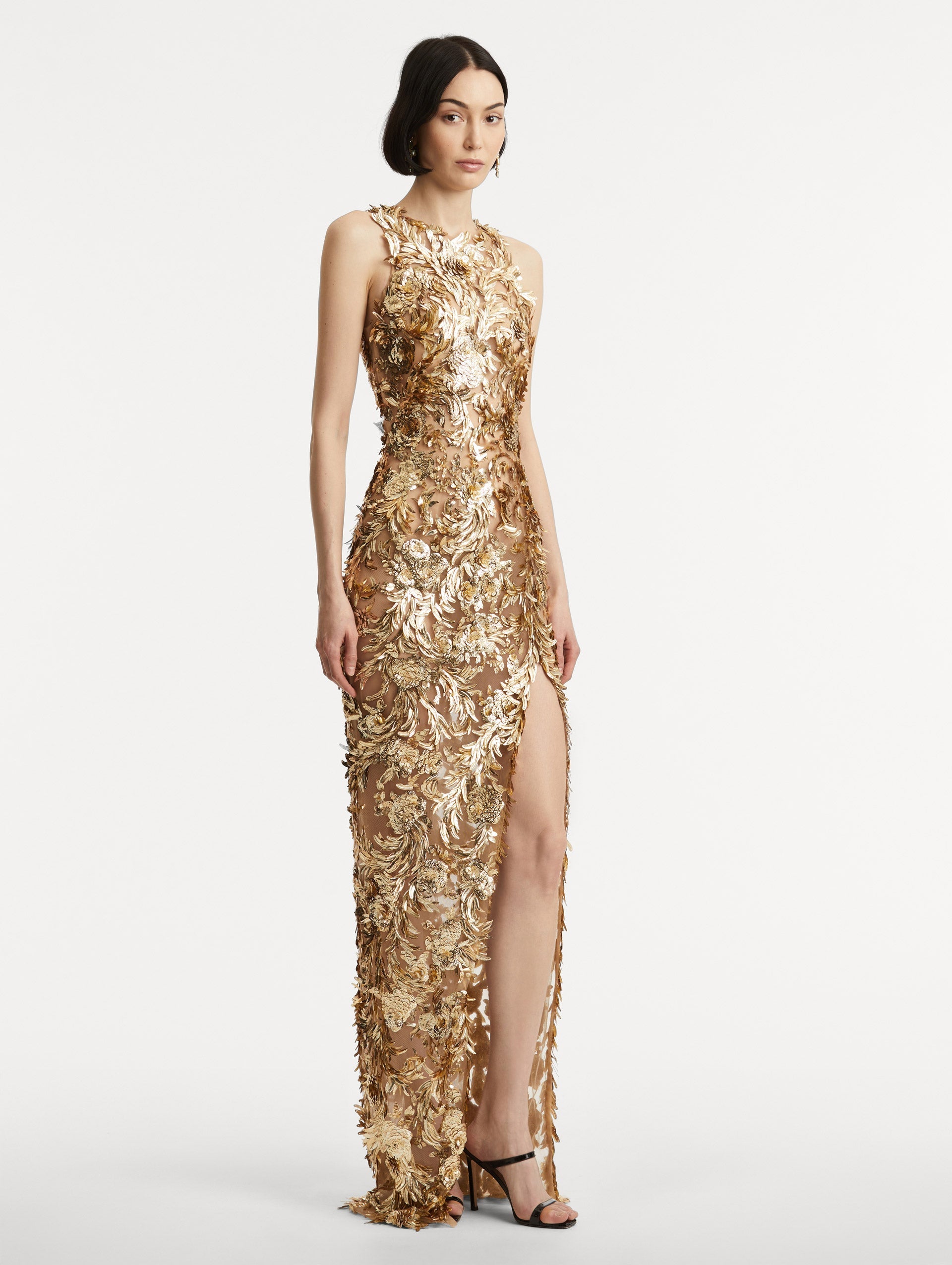 Full body side view of model wearing sleeveless gold-embroidered gown with a halter neckline and feathers unfurling across a tulle base. Gown features a high center front slit.