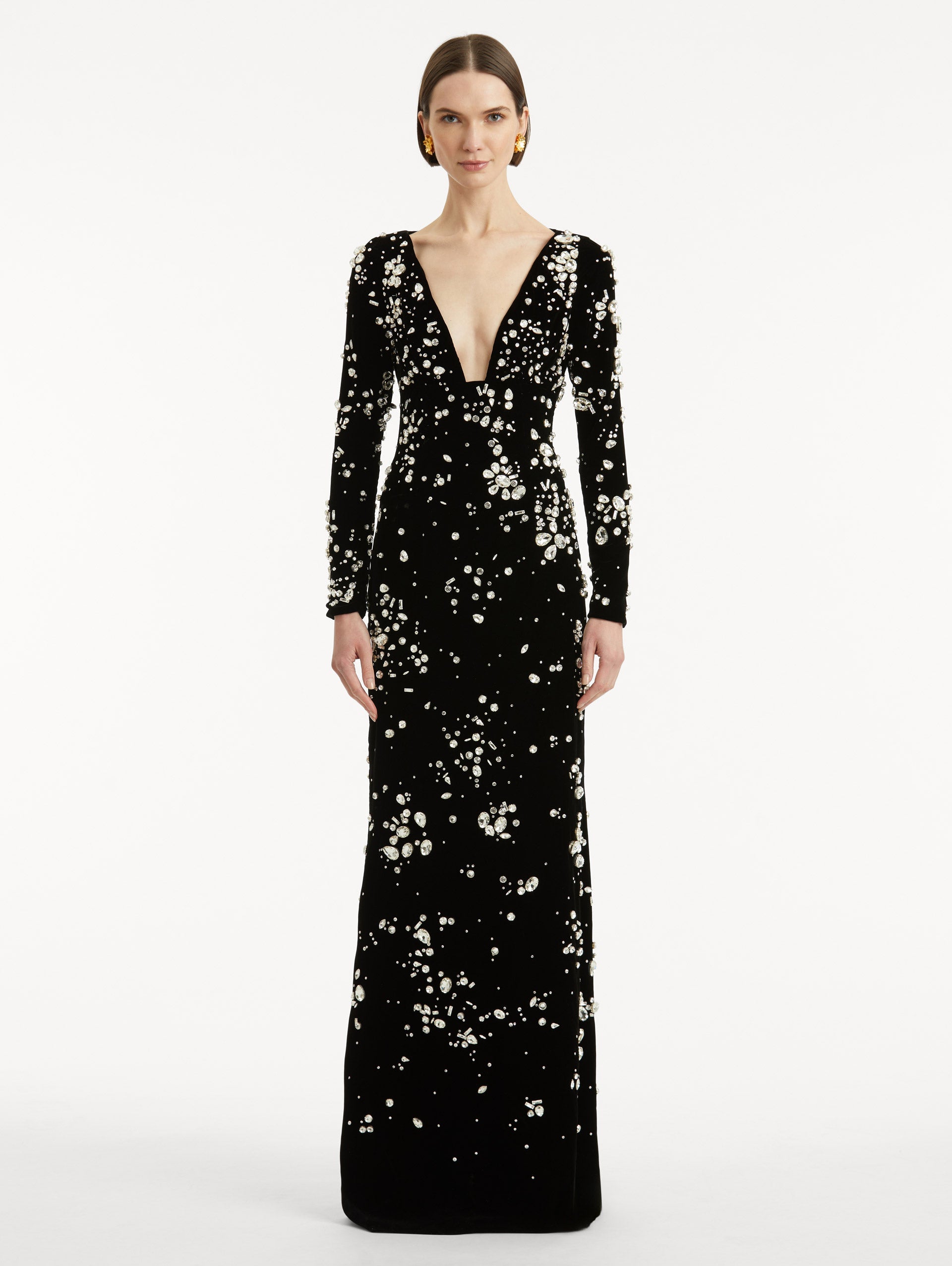 Full body front view of model wearing a long sleeve black gown with multi-dimensional stones scattered across the dress, features plunging V-neckline and column silhouette.
