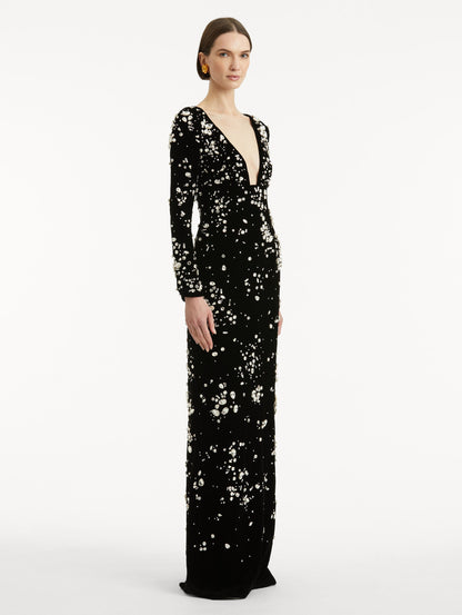 Full body side view of model wearing a long sleeve black gown with multi-dimensional stones scattered across the dress, features plunging V-neckline and column silhouette.