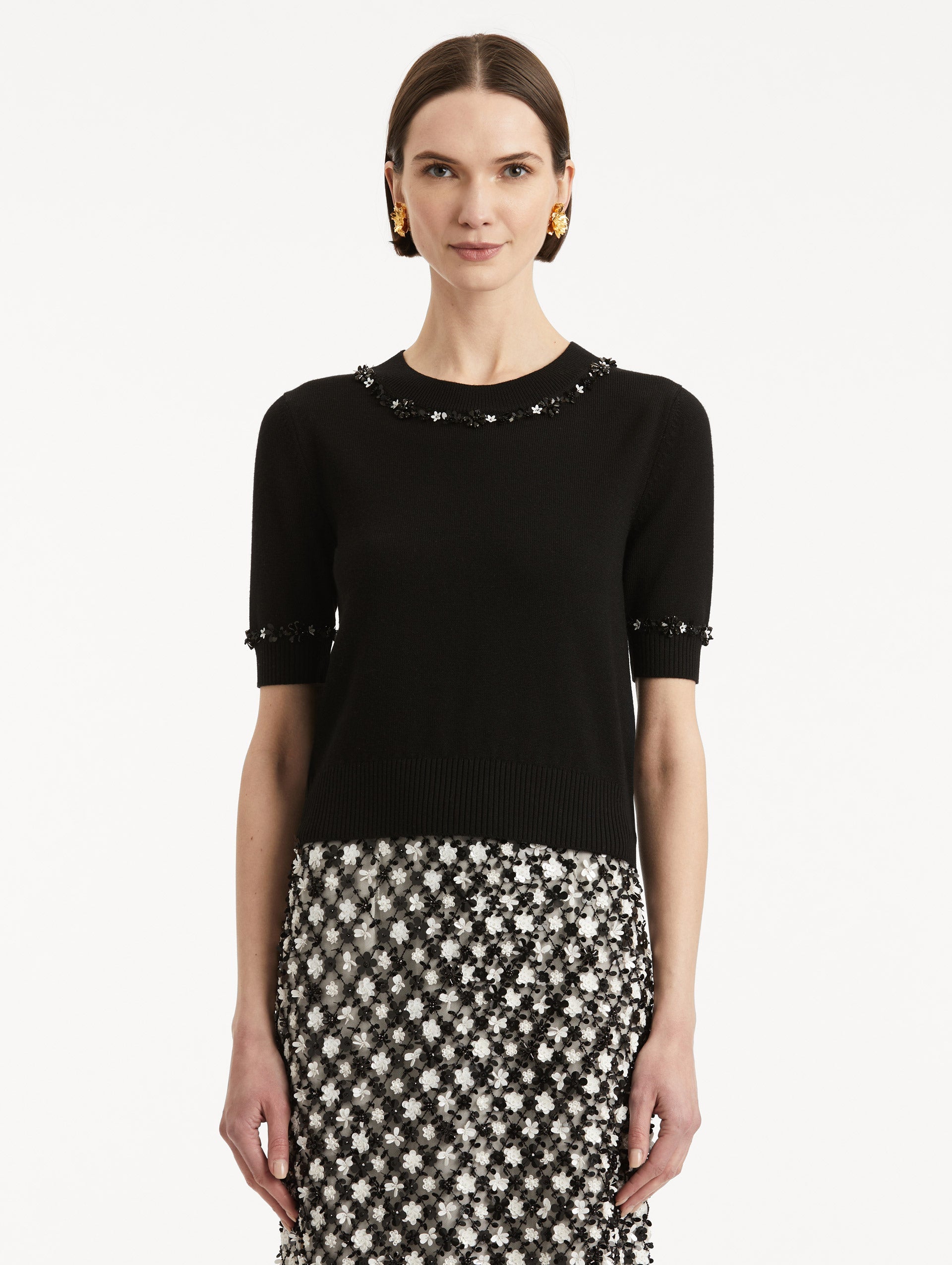 Front view of model wearing a short sleeve black pullover with a crew neckline and intricate sequin embroidery of ivory and noir florets which trace the trim.