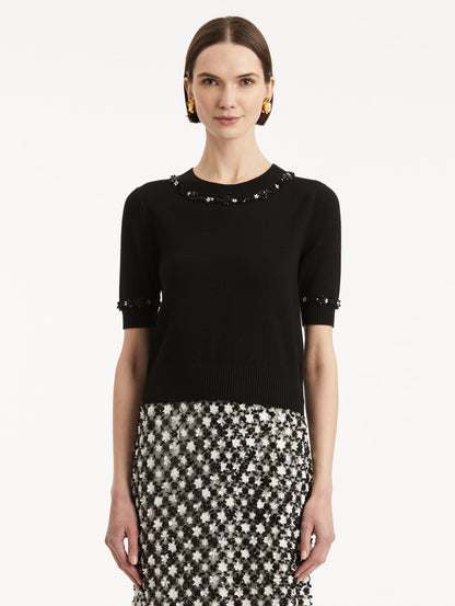 Front view of model wearing a short sleeve black pullover with a crew neckline and intricate sequin embroidery of ivory and noir florets which trace the trim.