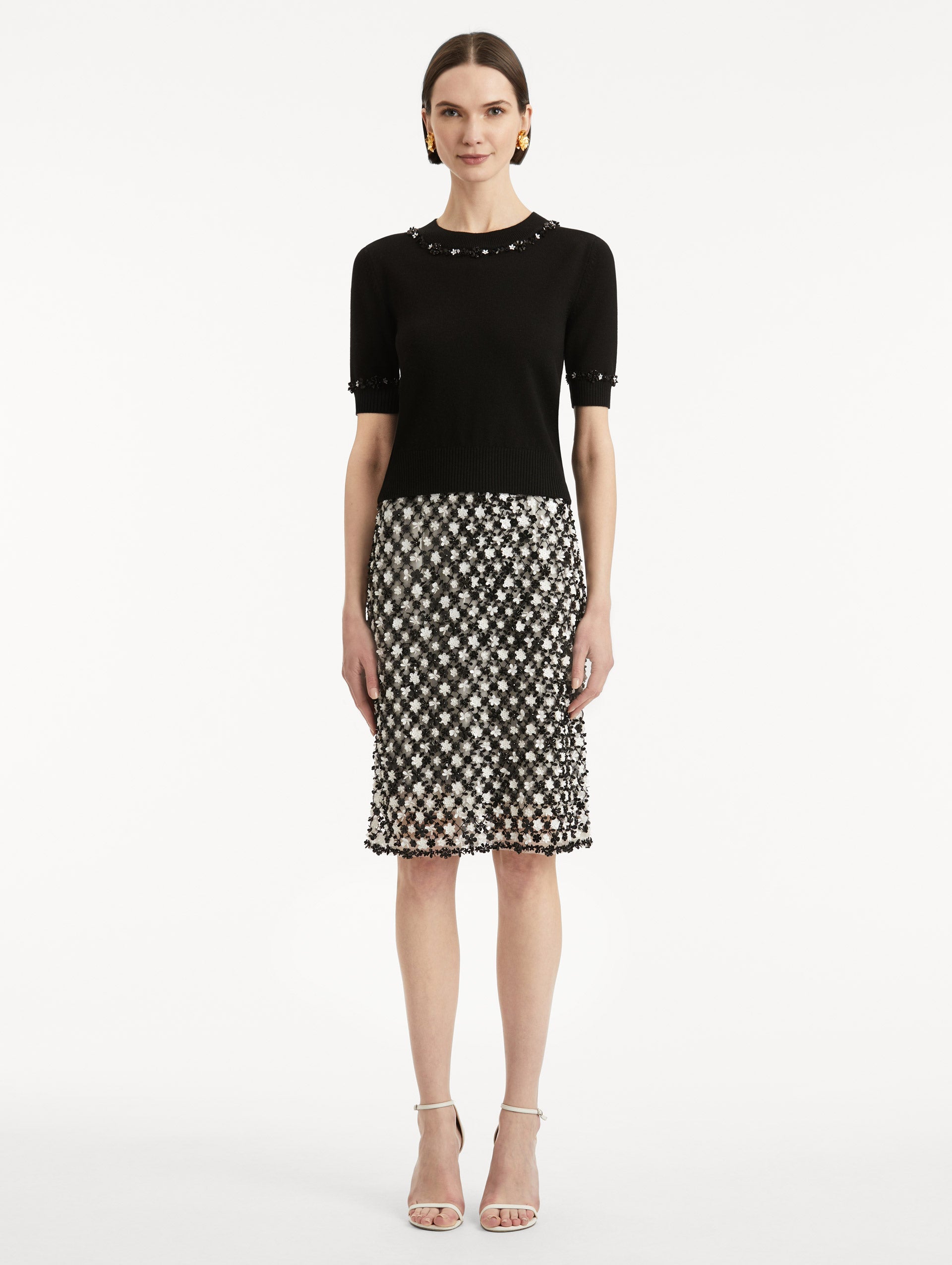 Full body front view of model wearing a short sleeve black pullover with a crew neckline and intricate sequin embroidery of ivory and noir florets which trace the trim