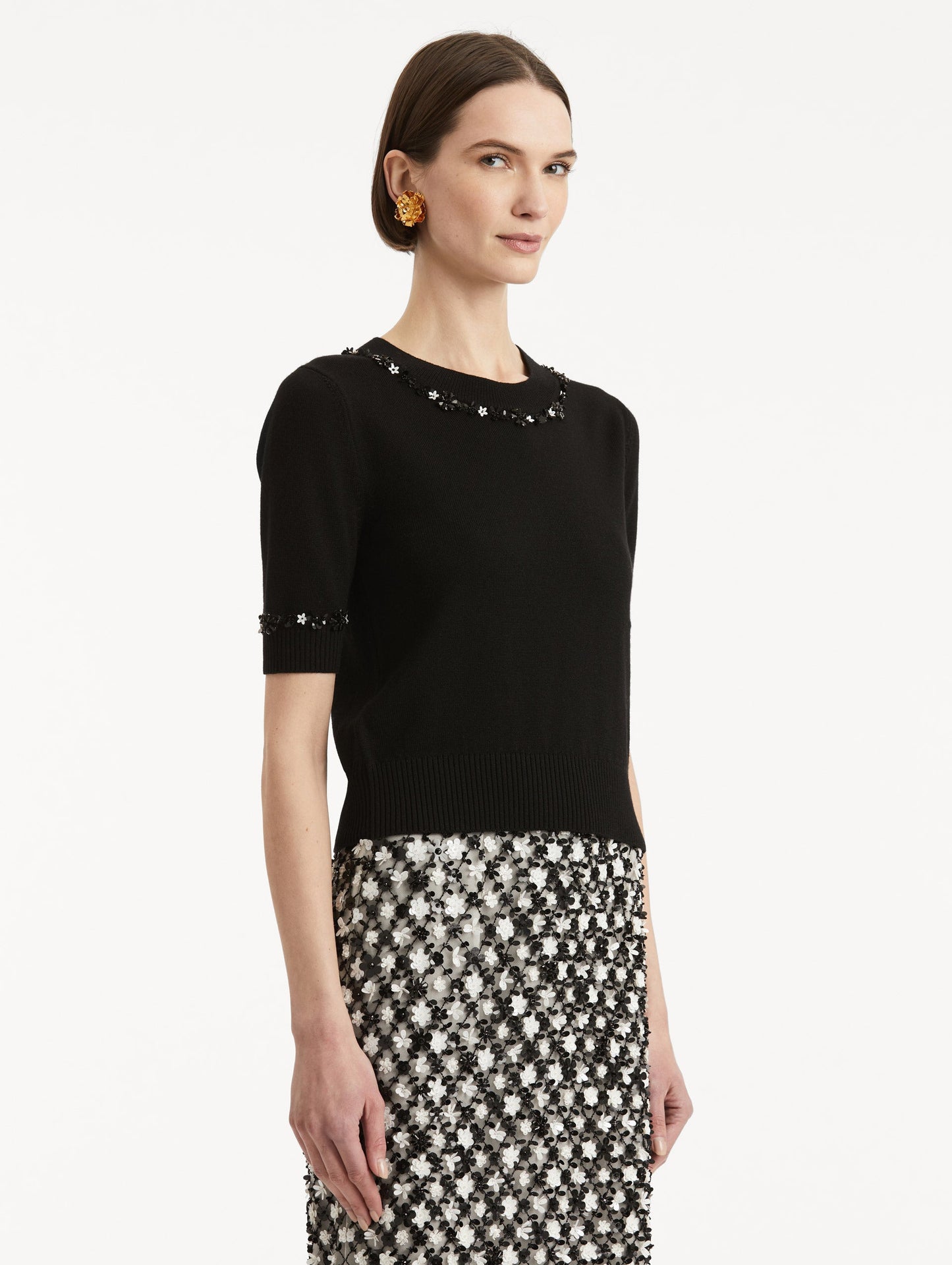 Side view of model wearing a short sleeve black pullover with a crew neckline and intricate sequin embroidery of ivory and noir florets which trace the trim