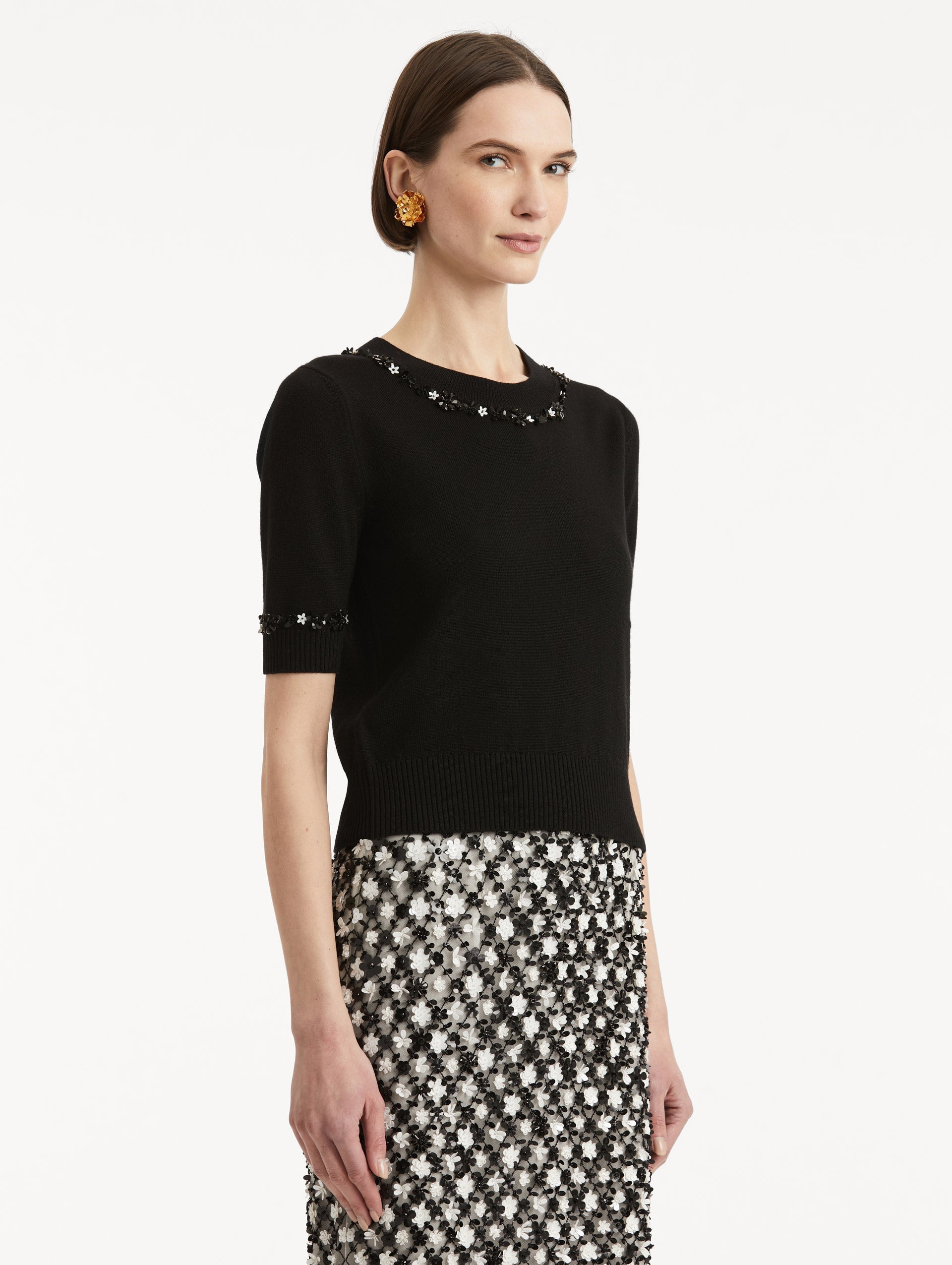 Side view of model wearing a short sleeve black pullover with a crew neckline and intricate sequin embroidery of ivory and noir florets which trace the trim
