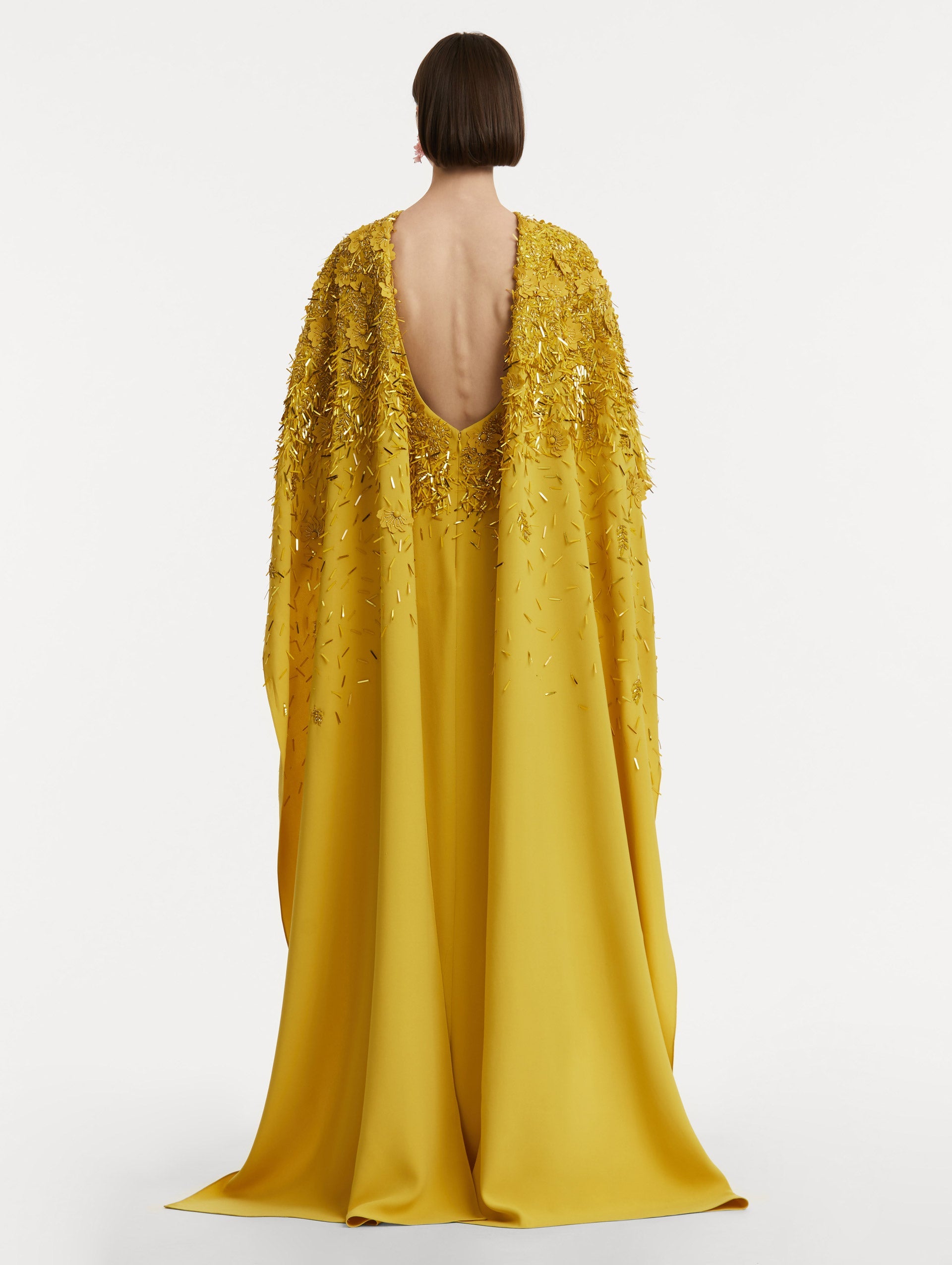 Full body back view of model wearing a yellow caftan with gold sequin and peony embroidery on draped caftan and surrounding the scoop back.