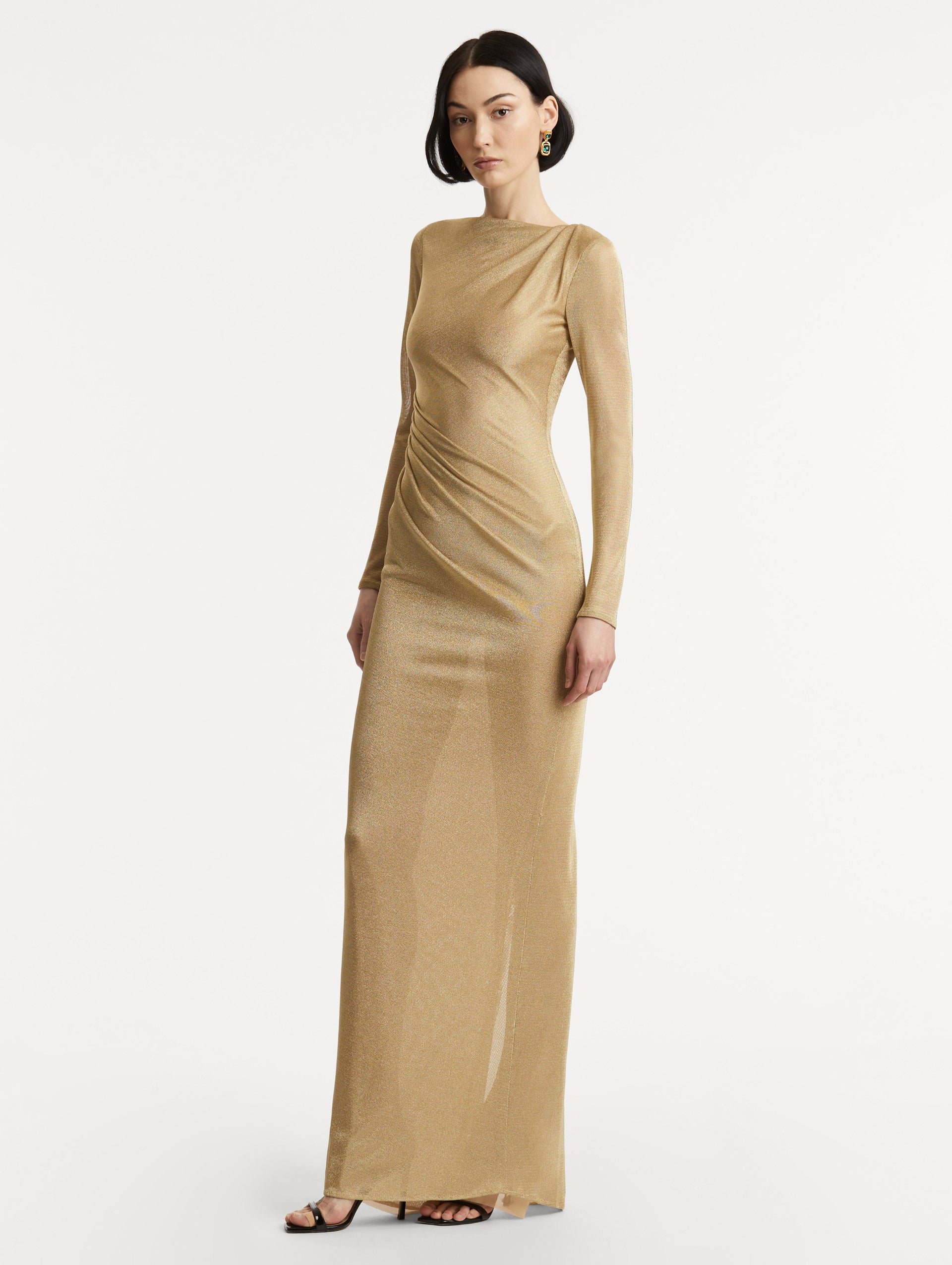 Full body side view image of model wearing a gold metallic jersey gown with a high neckline and long sleeves with draping at the shoulder and waist.