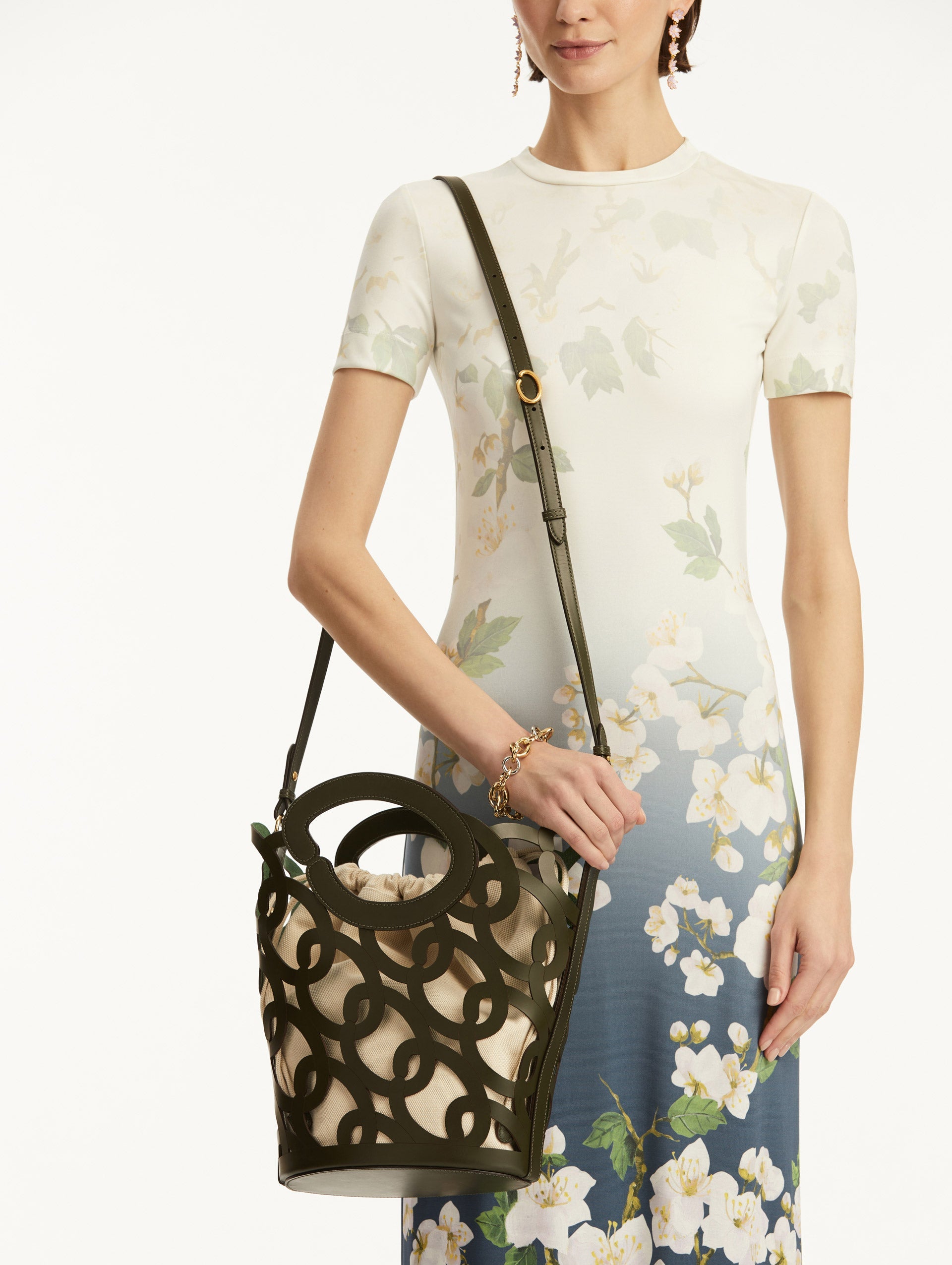 The model is holding the Oscar chain link bucket tote in olive green with a large o forming the top handle and detachable shoulder strap, paired with an ombre floral dress. Front View.