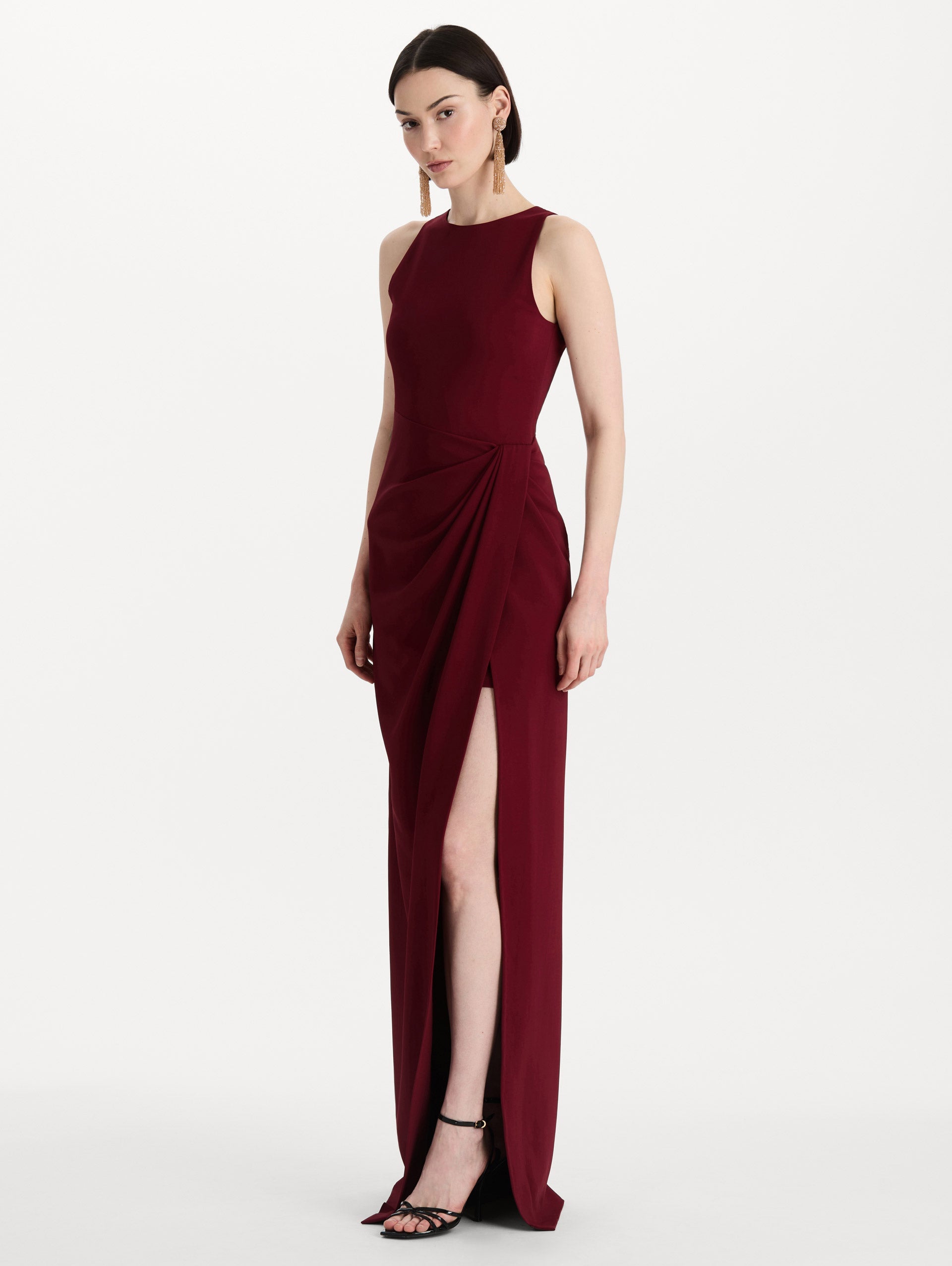 The model is wearing the drape-detailed cady gown with a sleeveless silhouette in the color claret. Side view.