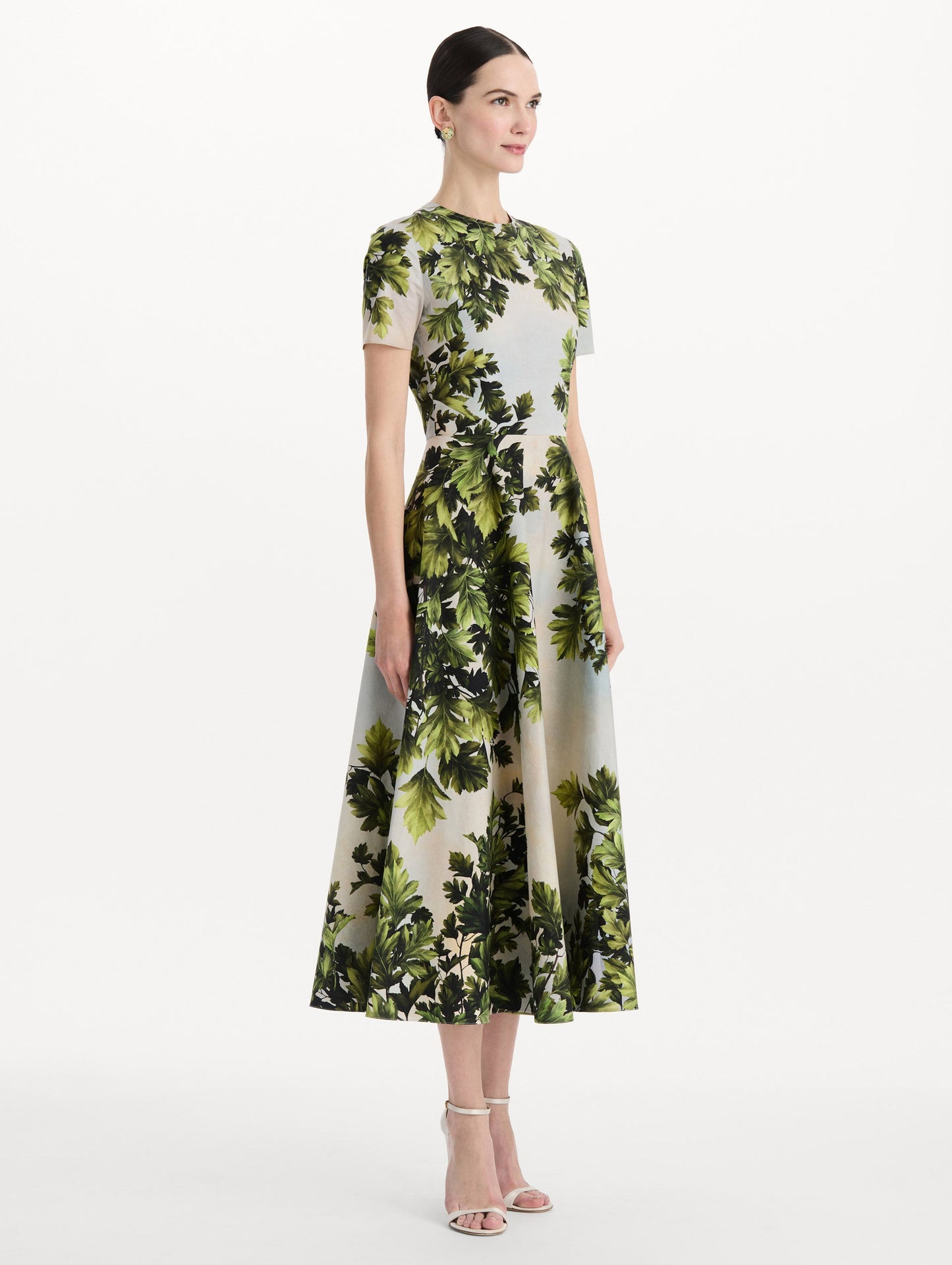 The model is wearing the maple leaves cotton poplin dress with short sleeves and an all-over foliage print in the color green multi. Side view.