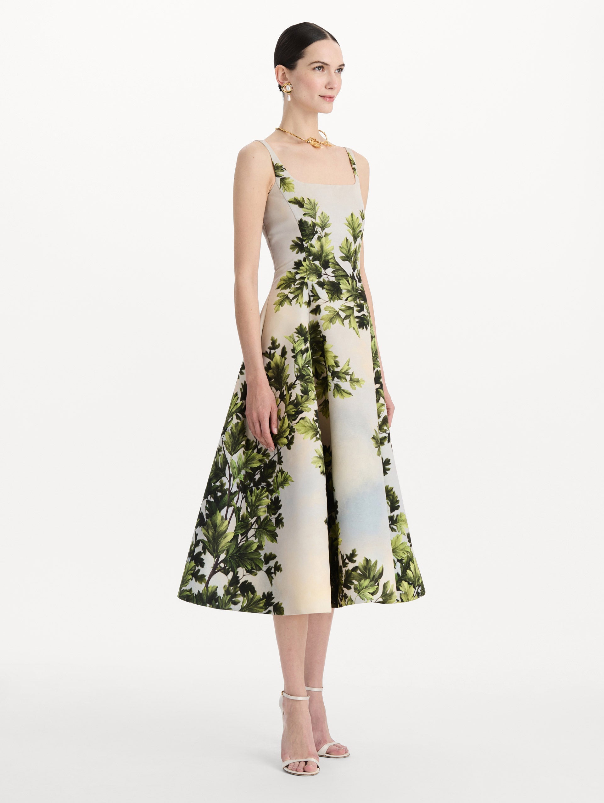 The model is wearing the maple leaves faille dress with a sleeveless scoop neckline and a foliage print in the color green multi. Side view.