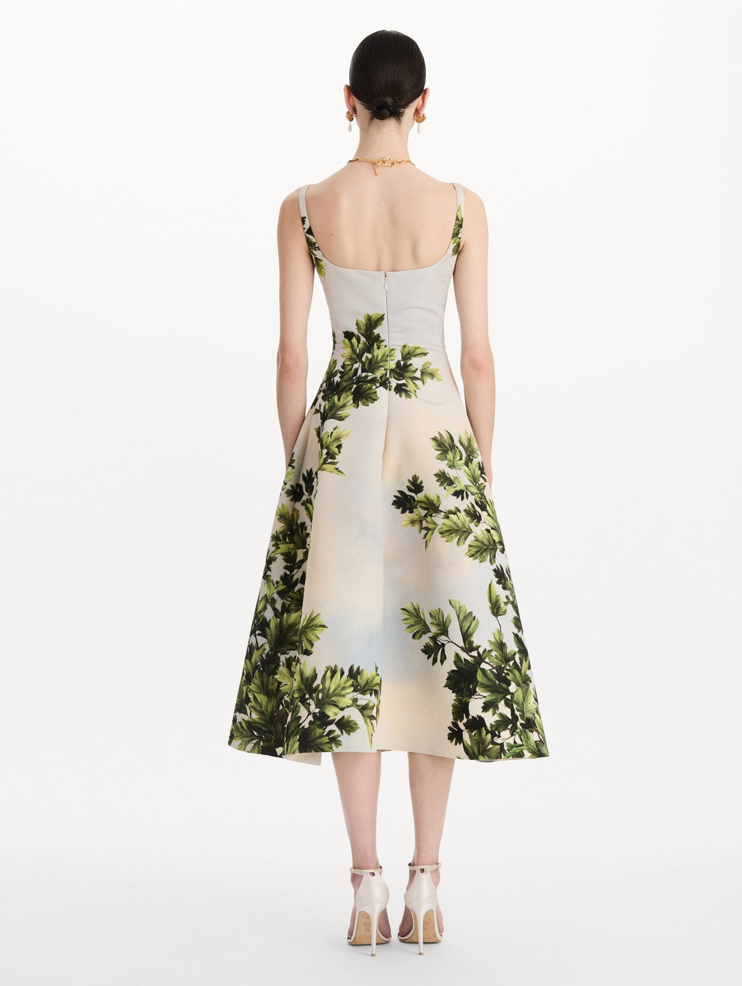 The model is wearing the maple leaves faille dress with a sleeveless scoop neckline and a foliage print in the color green multi. Back view.