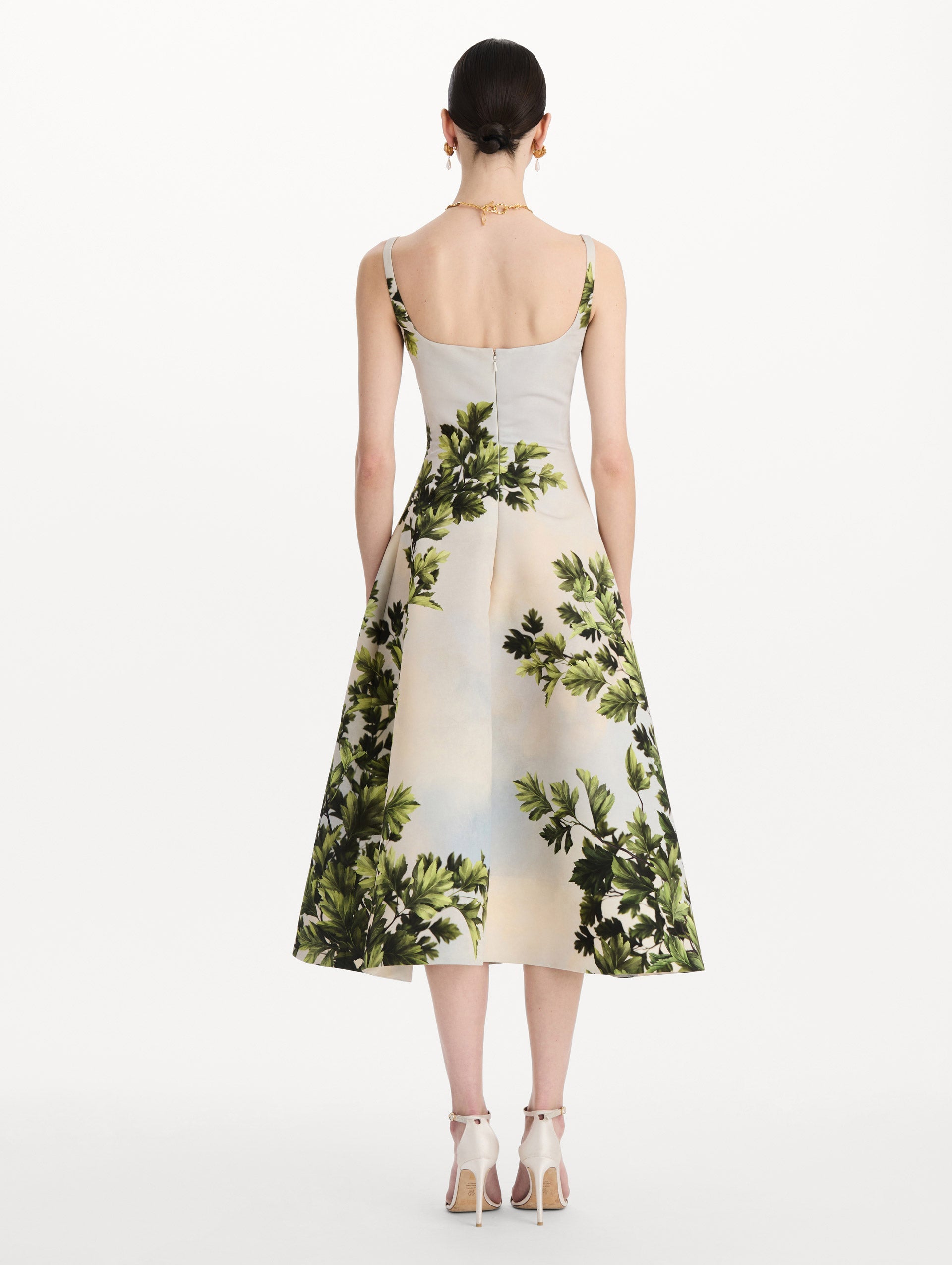 The model is wearing the maple leaves faille dress with a sleeveless scoop neckline and a foliage print in the color green multi. Back view.