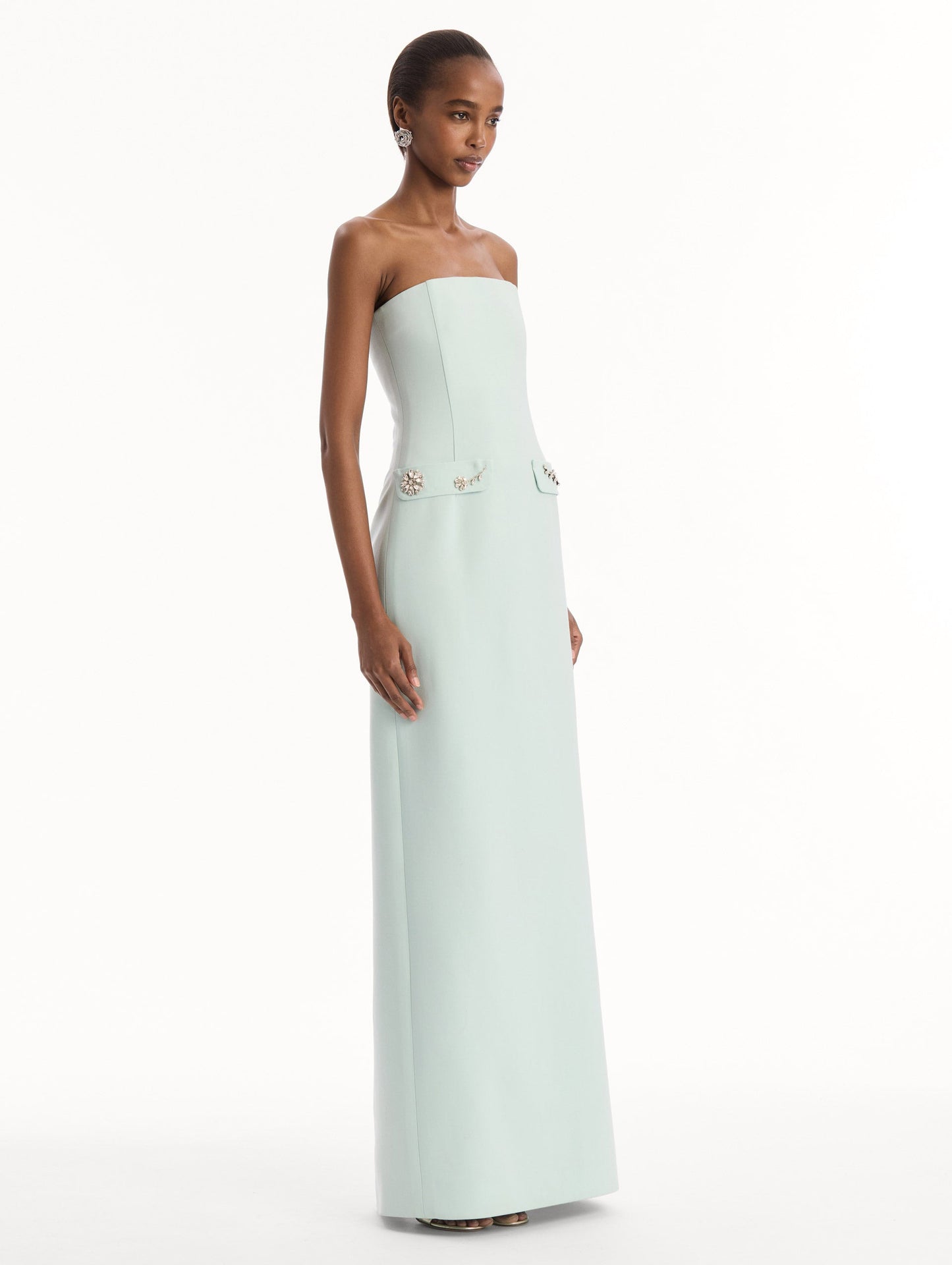 The model is wearing the Crystal Floral Embroidered Gown in Saltwater Blue. Featuring a strapless neckline, faux front flap pockets with crystal embroidery, and a long, column silhouette. Side View.
