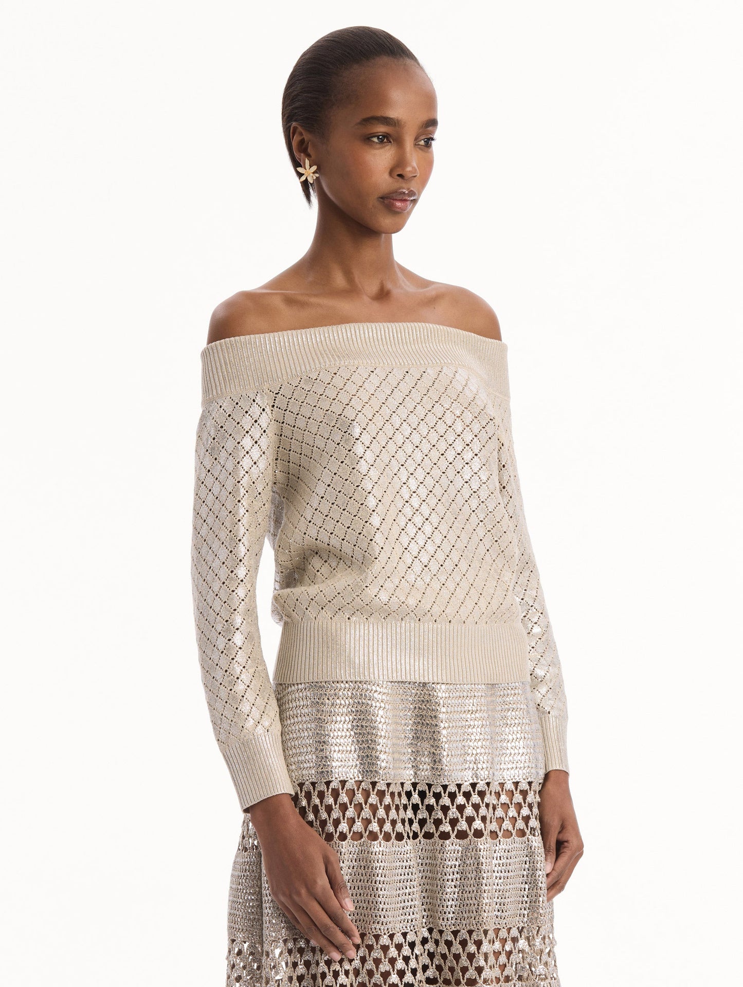 The model is wearing the Silver Foil Off-Shoulder Pullover. Featuring a lattice knit pattern, off-the-shoulder neckline, long sleeves, and ribbed trims. Side View.