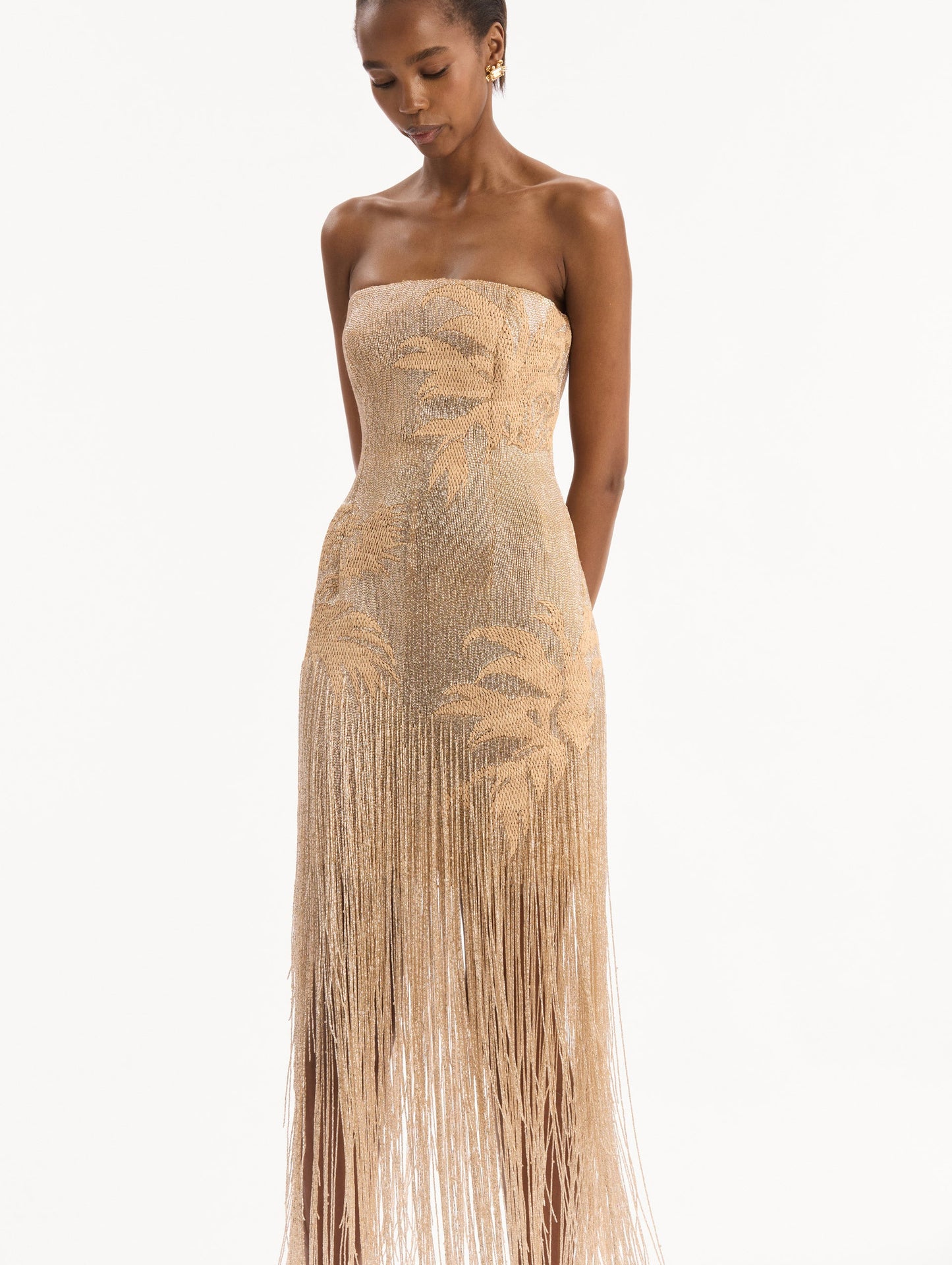 The model is wearing the Palm Tree Raffia Fringe Gown. Featuring a strapless neckline, raffia embroidery, and long, column silhouette with fringe skirt. Detail View.