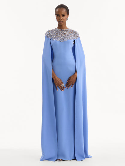 The model is wearing the Crystal & Glass Embroidered Caftan in a hydrangea blue color. Featuring crystal and glass bead embroidery at the collar, flowing cape-like sleeves, and a long skirt. Front View.