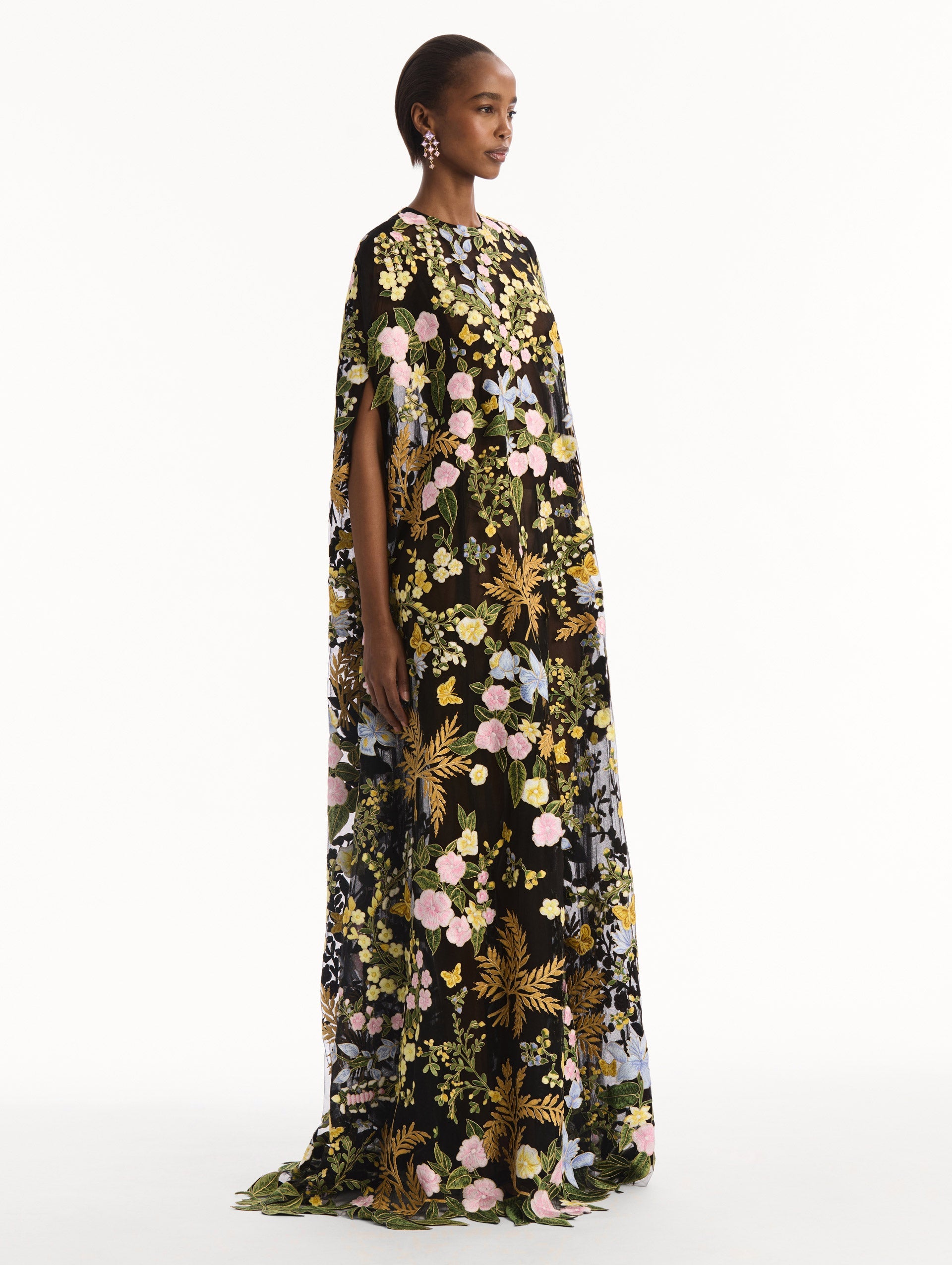 The model is wearing the Dominican Mixed-Floral Guipure Caftan. Featuring a long, flowing silhouette draping from the shoulders. Side View.