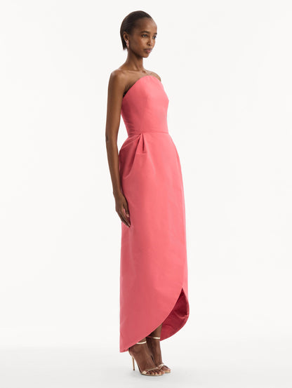 The model is wearing the Strapless Faille Gown in Guava Pink. Featuring a sculpted silhouette and wrap-style skirt with an asymmetrical hem. Side View.