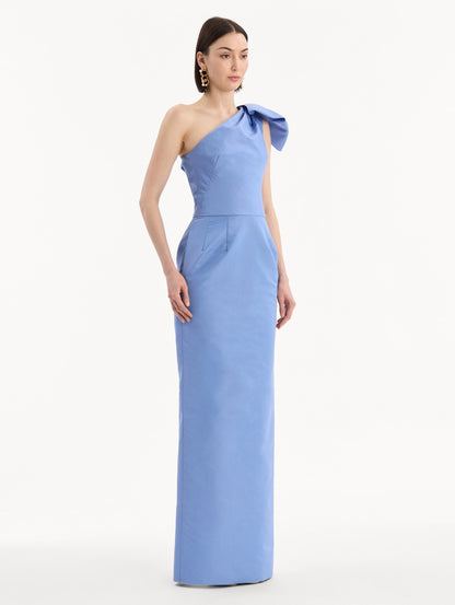 The model is wearing the Bow-Detail Faille Column Gown in a periwinkle blue. Featuring a column silhouette and sculptural bow detail at the shoulder. Side View.