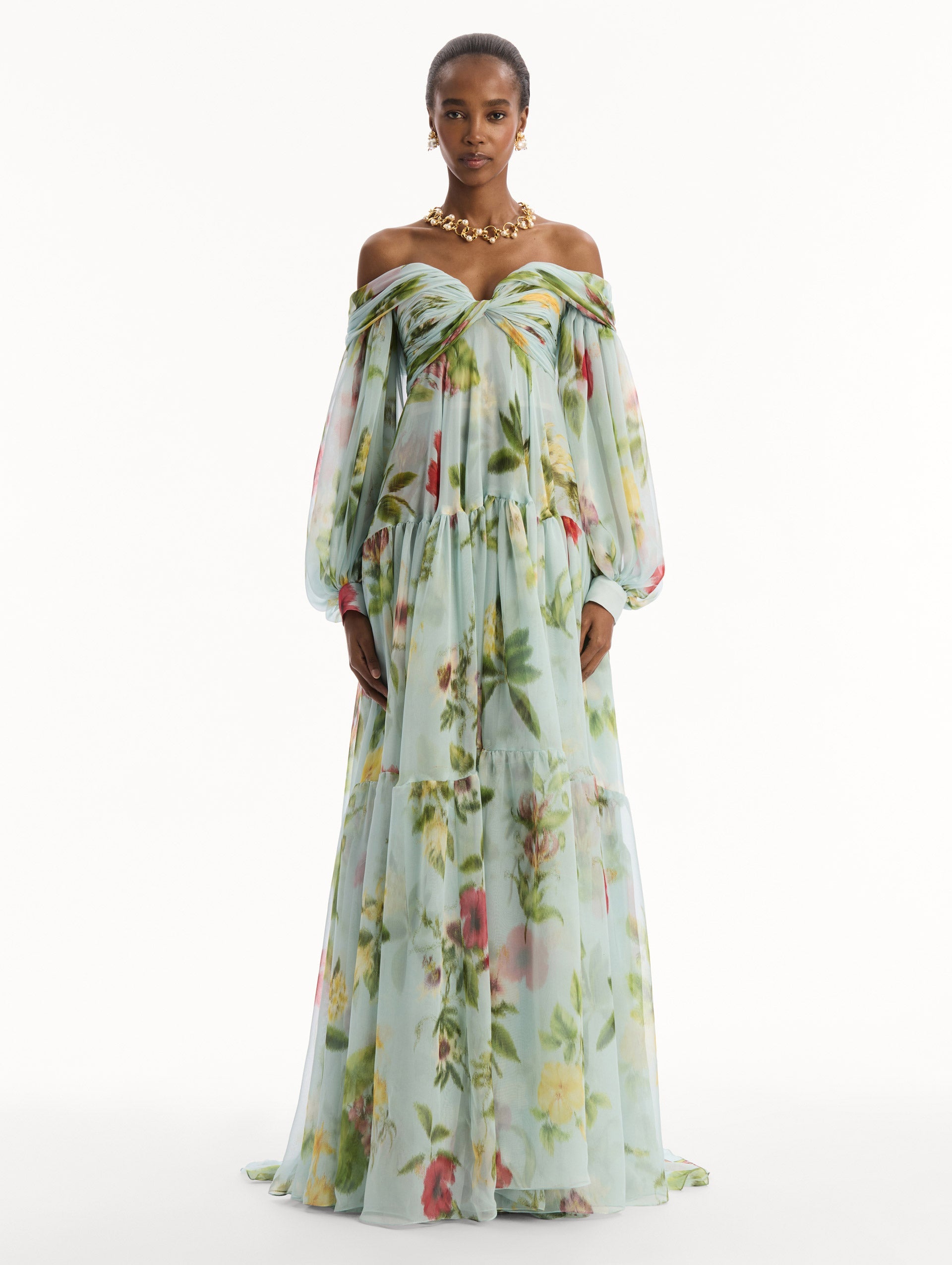 The model is wearing the Chiné Dominican Mixed-Floral Chiffon Sweetheart Gown. Featuring an off-the-shoulder sweetheart neckline, long sleeves, and flowing, tiered skirt. Front View.