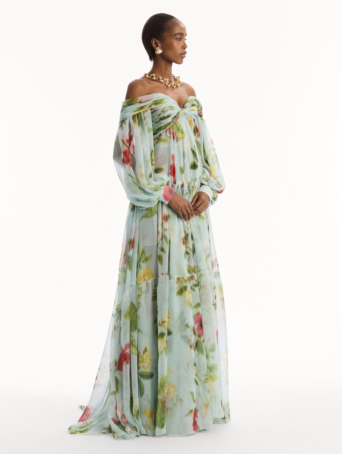 The model is wearing the Chiné Dominican Mixed-Floral Chiffon Sweetheart Gown. Featuring an off-the-shoulder sweetheart neckline, long sleeves, and flowing, tiered skirt. Side View.