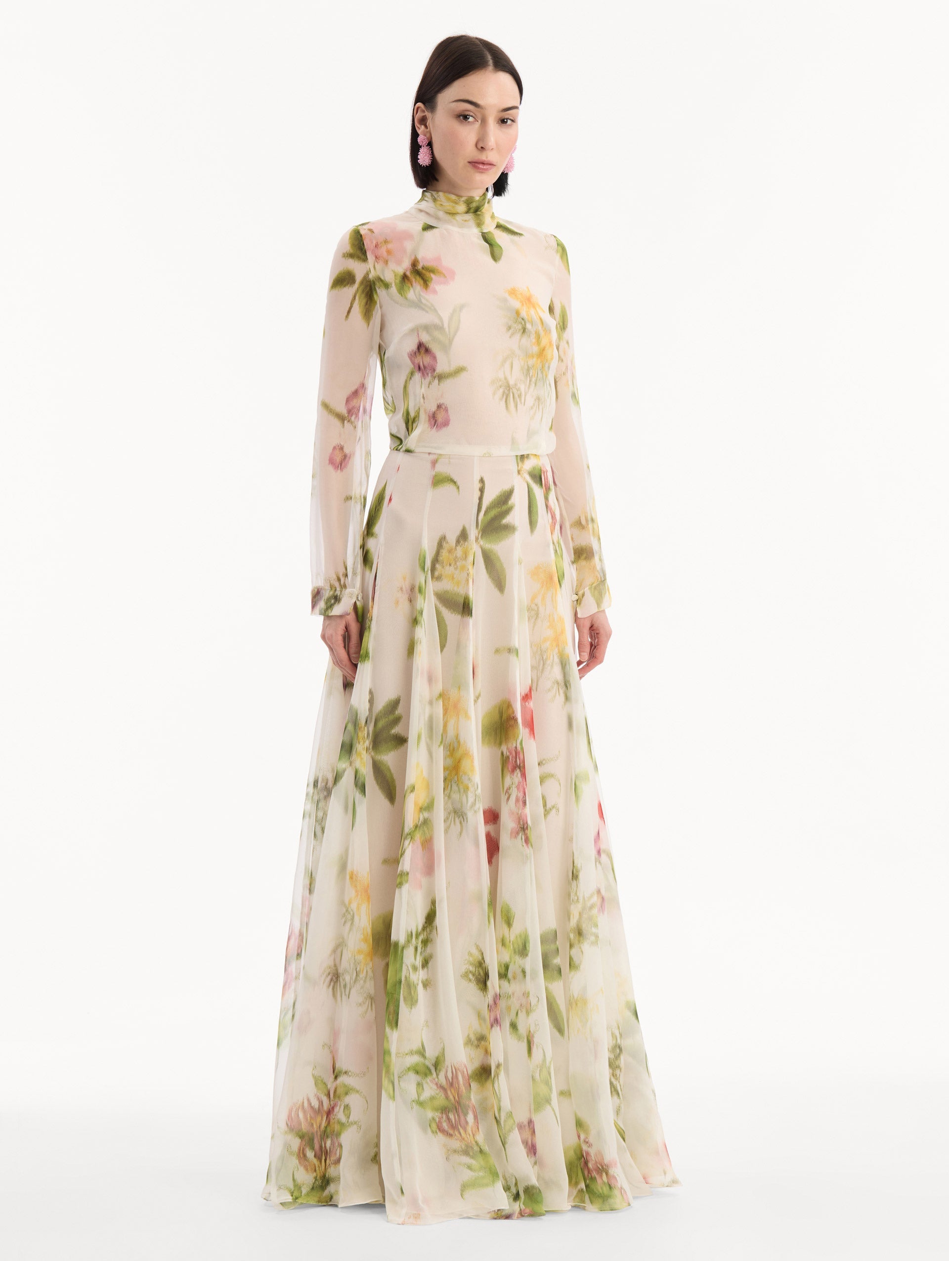 The model is wearing the Chiné Dominican Mixed-Floral Chiffon Gown in Ivory printed with an array of Dominican florals. Featuring long sleeves, mock neck, and flowing, pleated skirt. Side View.