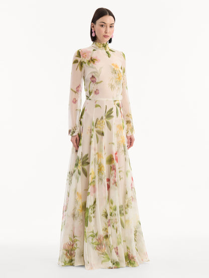 The model is wearing the Chiné Dominican Mixed-Floral Chiffon Gown in Ivory printed with an array of Dominican florals. Featuring long sleeves, mock neck, and flowing, pleated skirt. Side View.