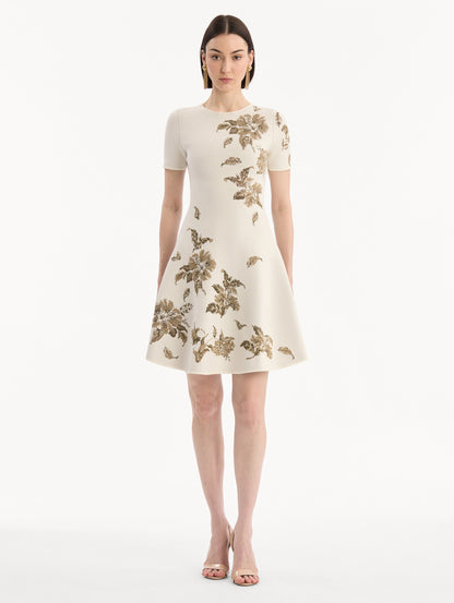The model is wearing the Hibiscus Metallic Jacquard Dress in Ivory with Gold hibiscus motifs. Featuring short sleeves, a softly structured silhouette, and mini-length skirt. Front View.
