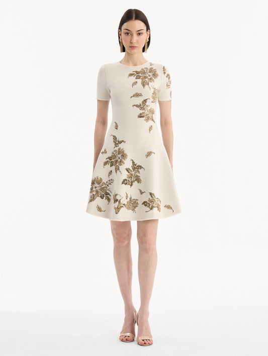 The model is wearing the Hibiscus Metallic Jacquard Dress in Ivory with Gold hibiscus motifs. Featuring short sleeves, a softly structured silhouette, and mini-length skirt. Front View.