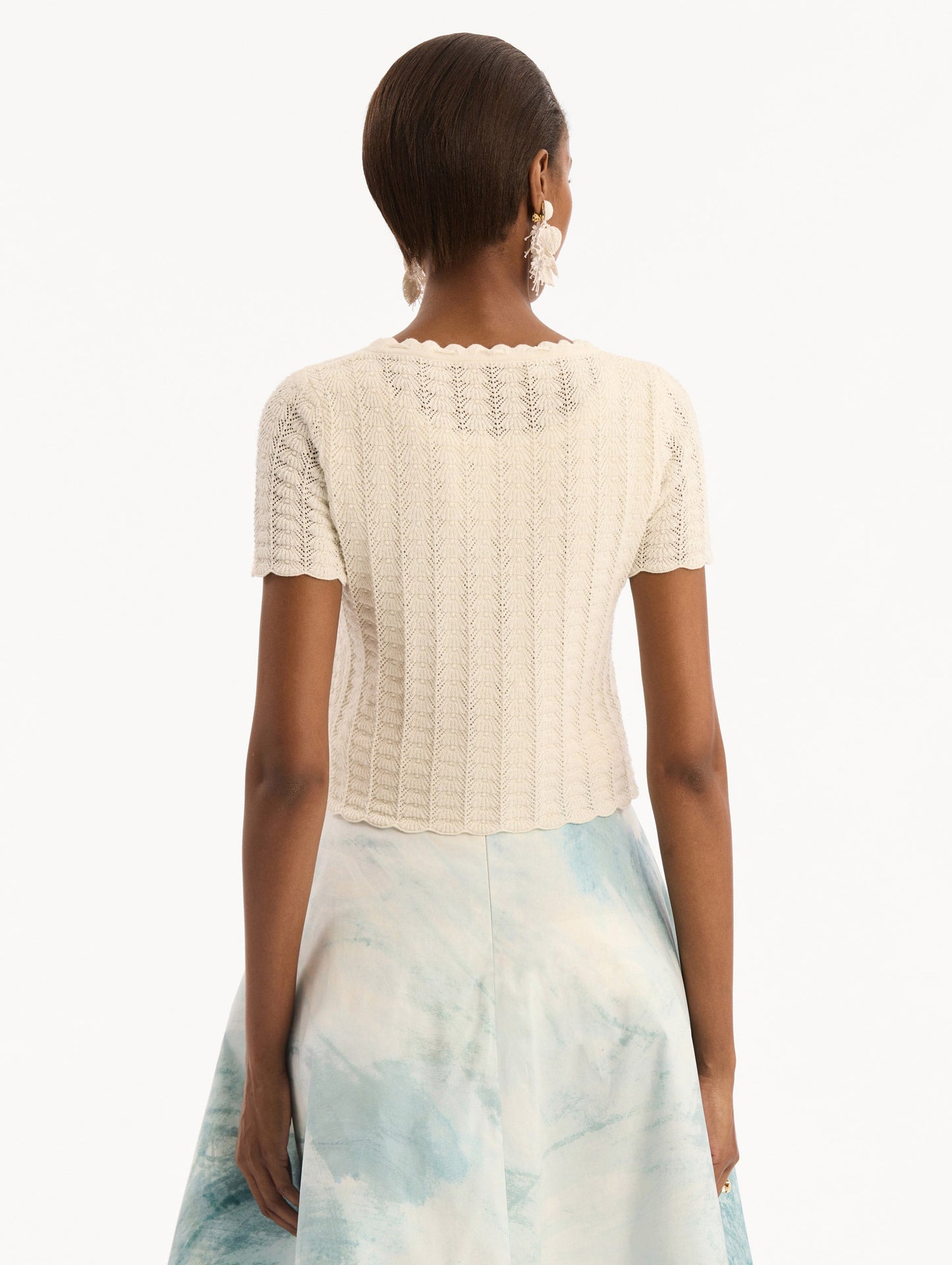 The model is wearing the Pointelle Weave Cardigan in Ivory. Featuring a scoop neckline, short sleeves, scalloped edges, and a front tie closure. Back View.