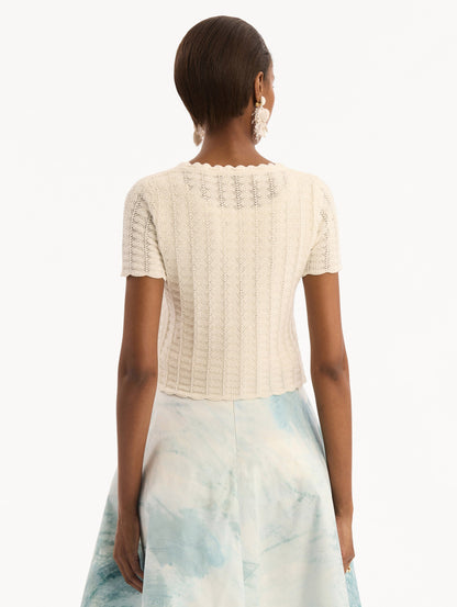 The model is wearing the Pointelle Weave Cardigan in Ivory. Featuring a scoop neckline, short sleeves, scalloped edges, and a front tie closure. Back View.