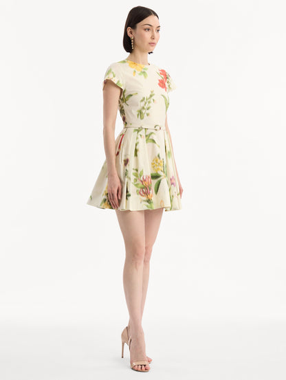 The model is wearing the Chiné Dominican Mixed-Floral Cotton Poplin Dress in Ivory printed with an array of Dominican florals. Featuring cap sleeves, belt at waist, and gathered mini skirt. Side View.