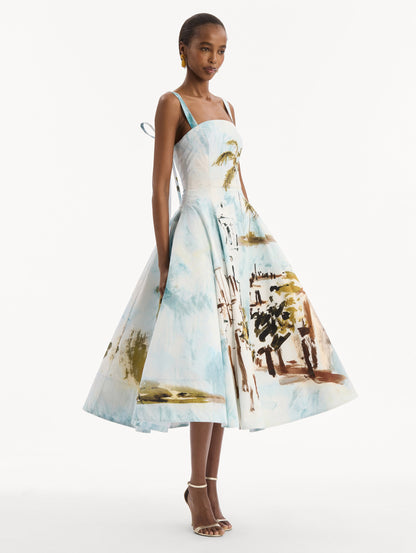 The model is wearing the Dominican Landscape Cotton Poplin Sleeveless Dress. Featuring tank straps with bow feature in back, and a full, midi-length skirt. Side View.