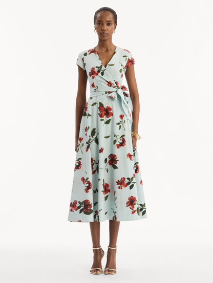 The model is wearing the Hibiscus Cotton Poplin Wrap Dress in light blue printed with red hibiscus blooms. Featuring a wrap construction bodice, cap sleeves, V-neckline, and full midi skirt. Front View.