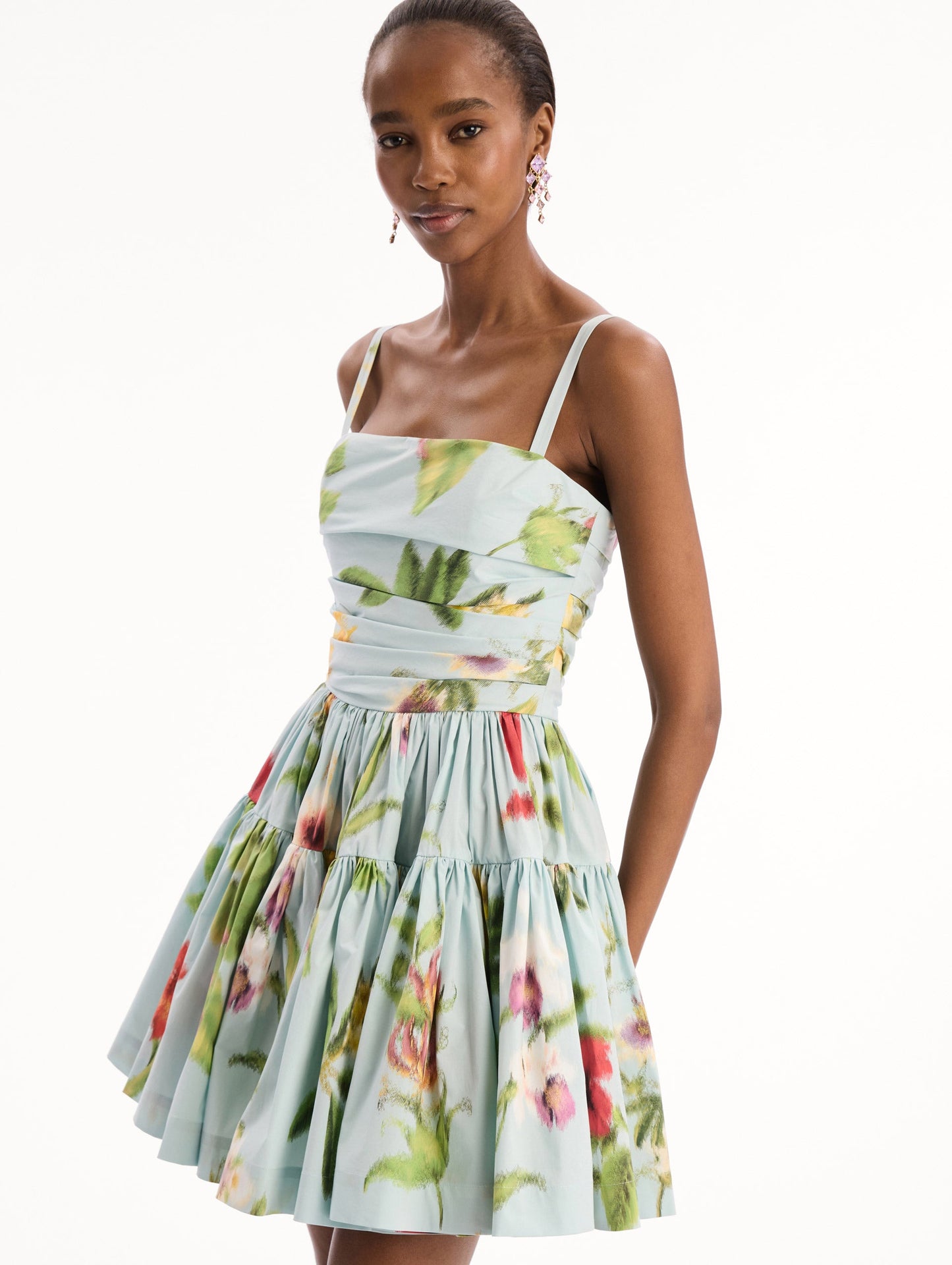 The model is wearing the Chiné Dominican Mixed-Floral Cotton Poplin Tiered Dress in light blue printed with a vibrant array of Dominican florals. Featuring a ruched bodice, tank straps, and gathered mini skirt. Detail View.