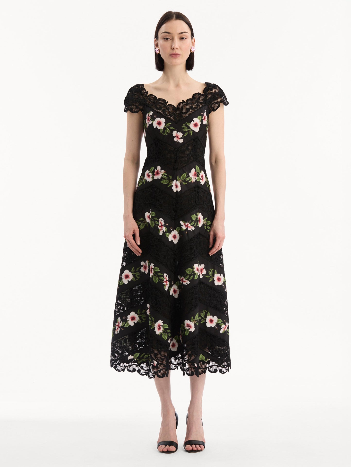 The model is wearing the Chiné Hibiscus Striped Faille Midi Dress in Black. Featuring off-the-shoulder cap sleeves, a midi length, and woven hibiscus motifs alternating with panels of faille and guipure lace, creating a striped composition. Front View.