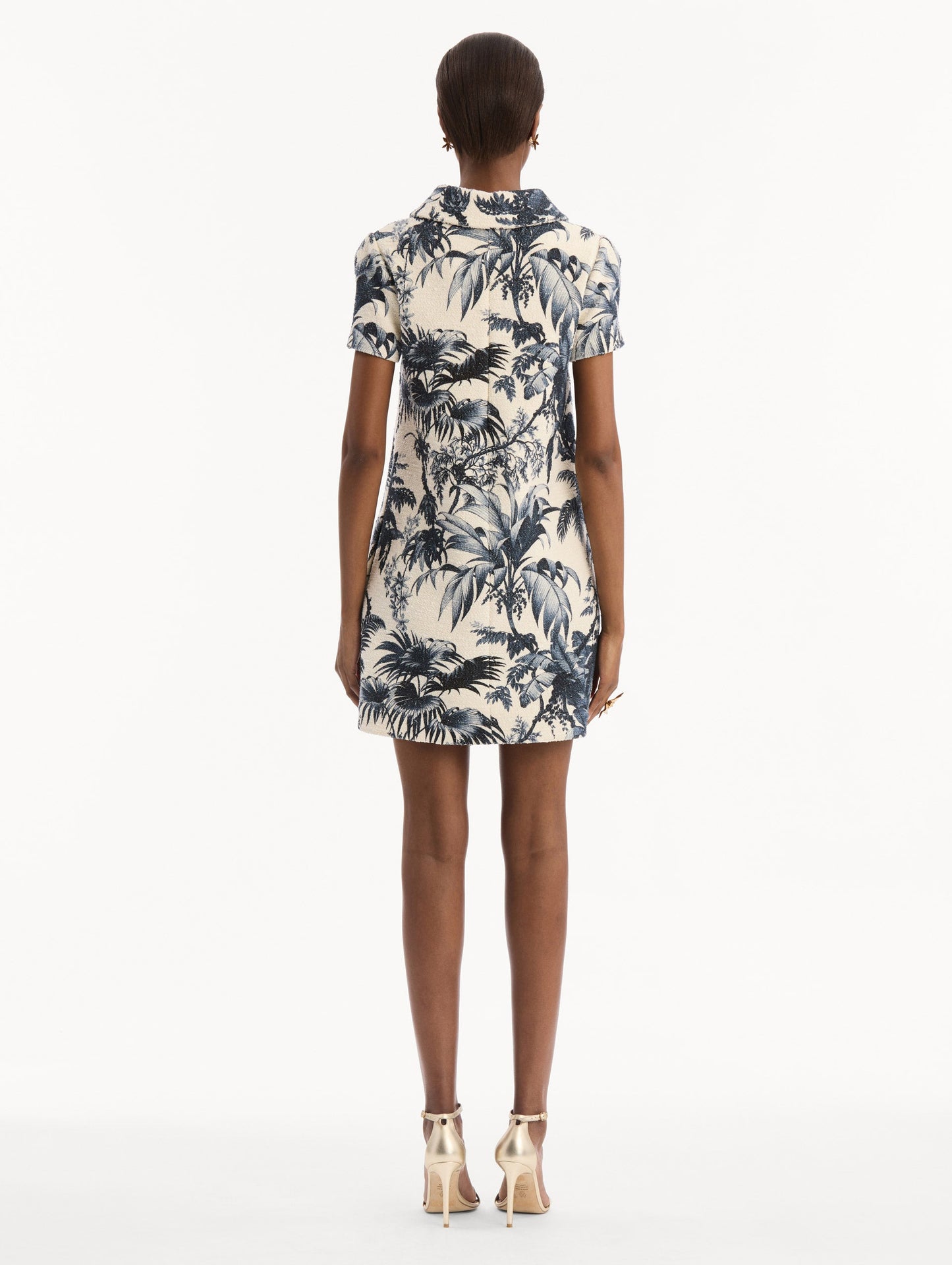 The model is wearing the Chiné Palm Leaves Shift Dress in ivory printed with navy palm leaves. Featuring a classic collar neckline, short sleeves, and mini length. Back View.