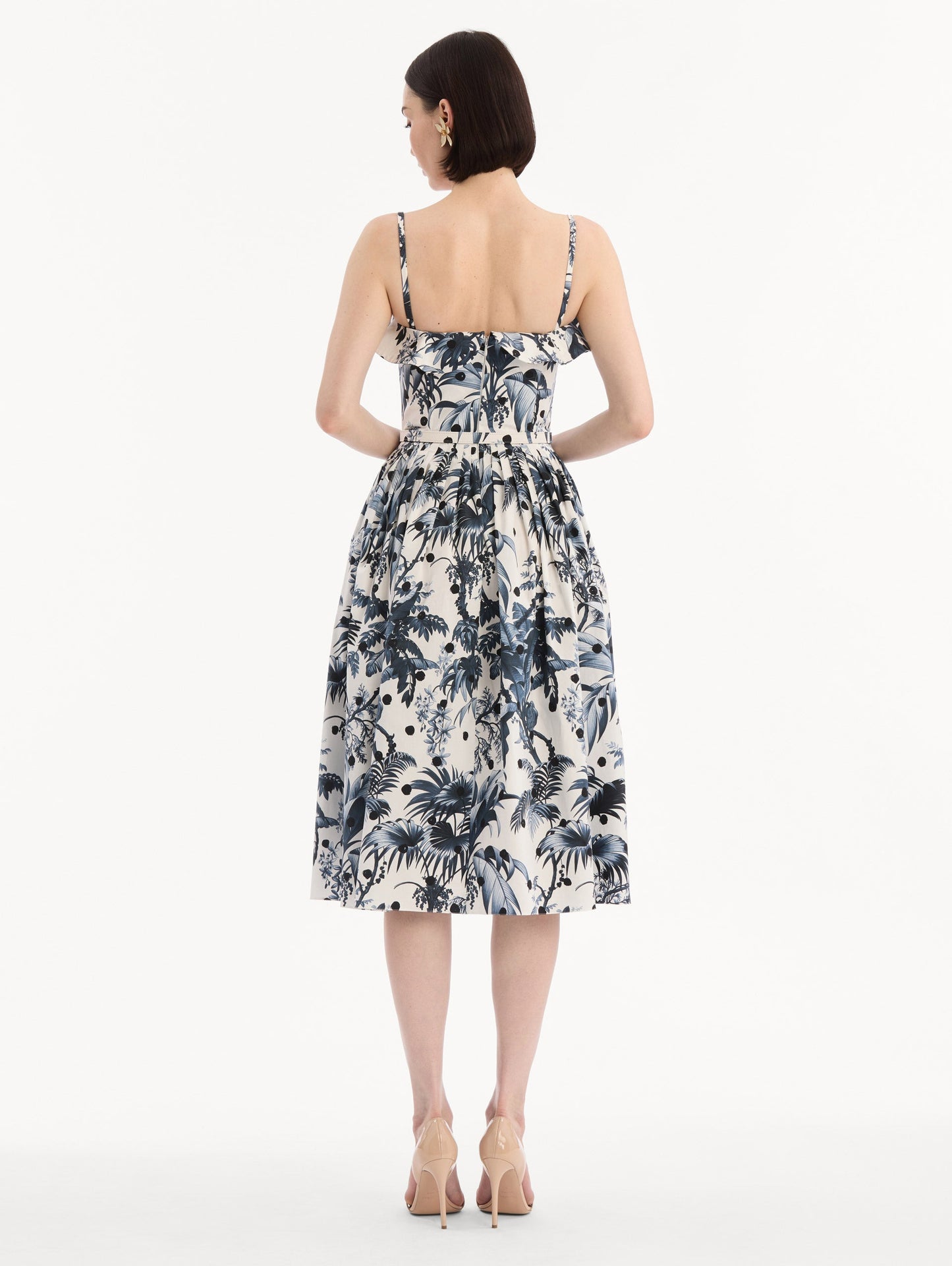 The model is wearing the Dotted Palm Leaves Cotton Poplin Sleeveless Dress in Navy Blue and White. Featuring a palm leaves print, ruffled top, delicate straps, belt at waist, softly structured bodice, and gathered skirt. Back View.